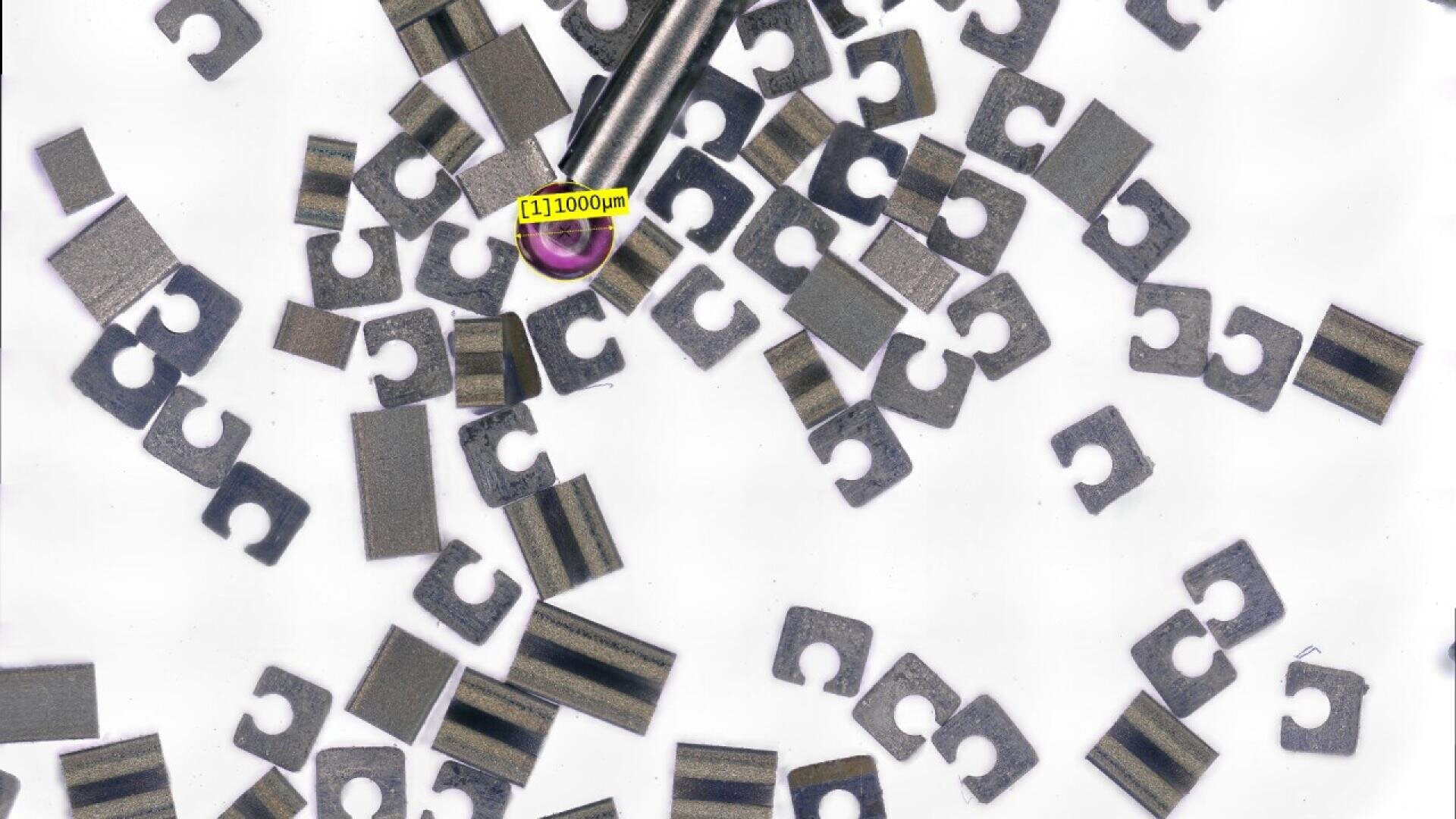 A close-up image shows various tiny metallic micro-components scattered on a white surface, with a tweezer holding one piece near a yellow scale label marked 1000 µm.