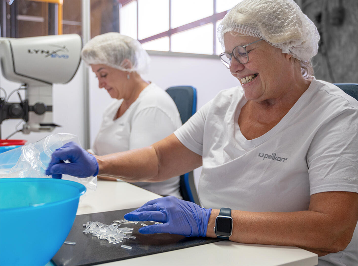 Two women wearing white shirts, hairnets, and gloves sit at a table handling small clear plastic parts. One woman is smiling while working; a microscope and blue bowl are also visible on the table.