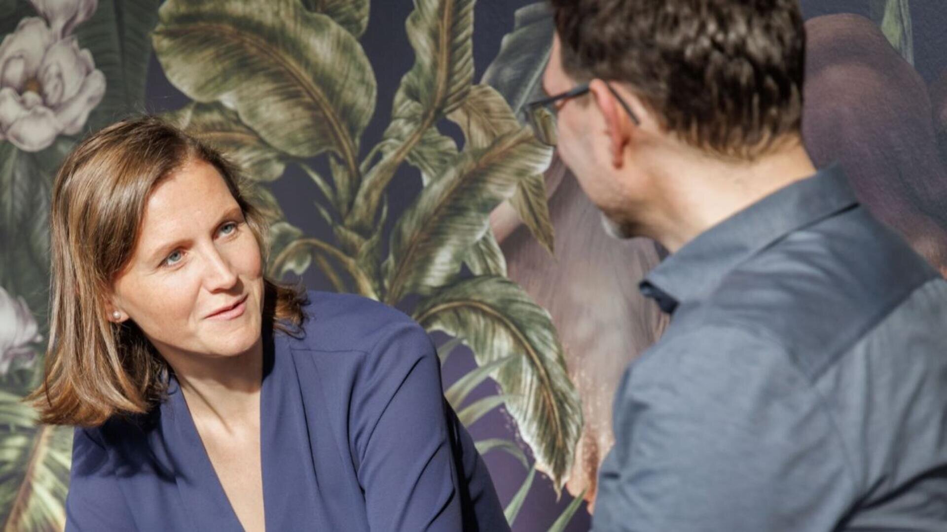 A woman in a blue blouse attentively listens to a man wearing glasses and a blue shirt, with a leafy, nature-inspired background behind them.