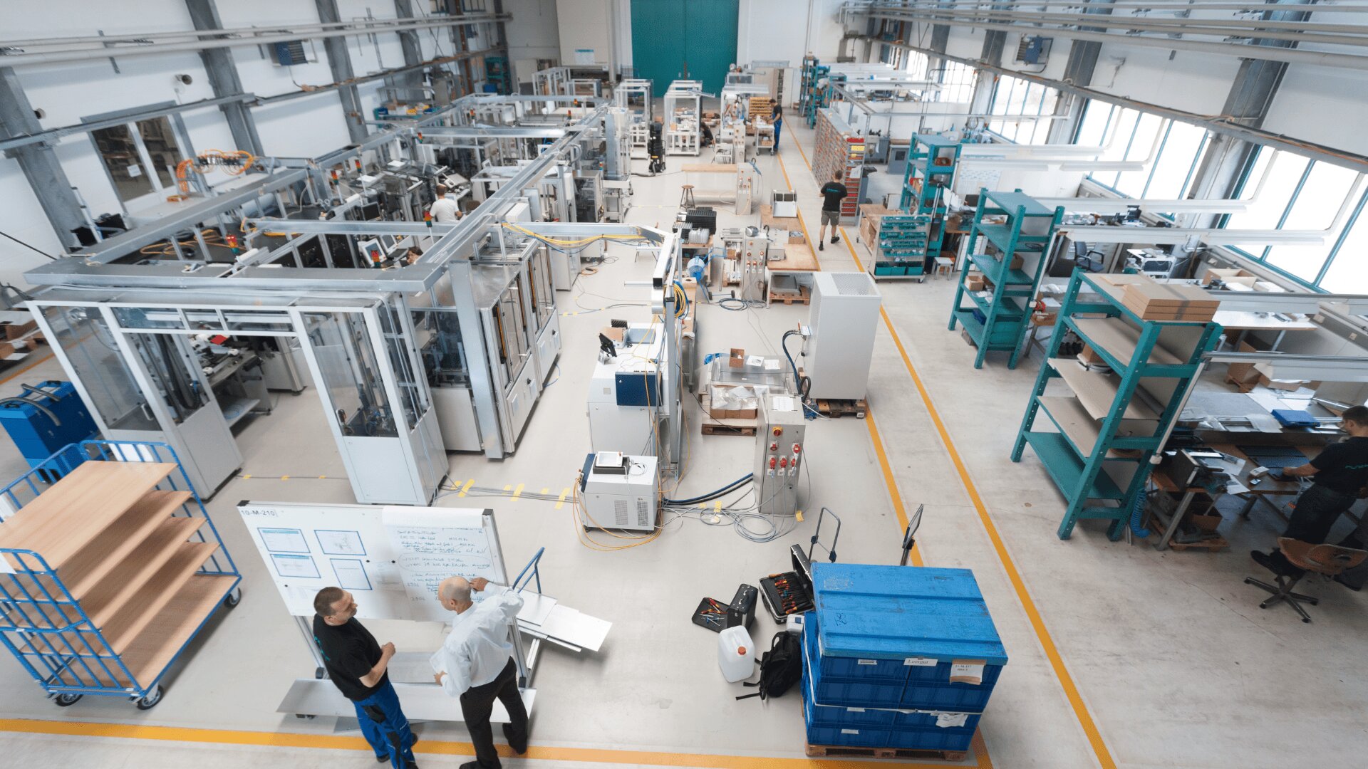 Draufsicht auf eine geräumige, moderne Fabrik mit Maschinen, Regalen, Arbeitsplätzen und zwei Personen im Gespräch auf der linken Seite. Der Arbeitsbereich ist übersichtlich gestaltet und durch große Fenster hell erleuchtet.