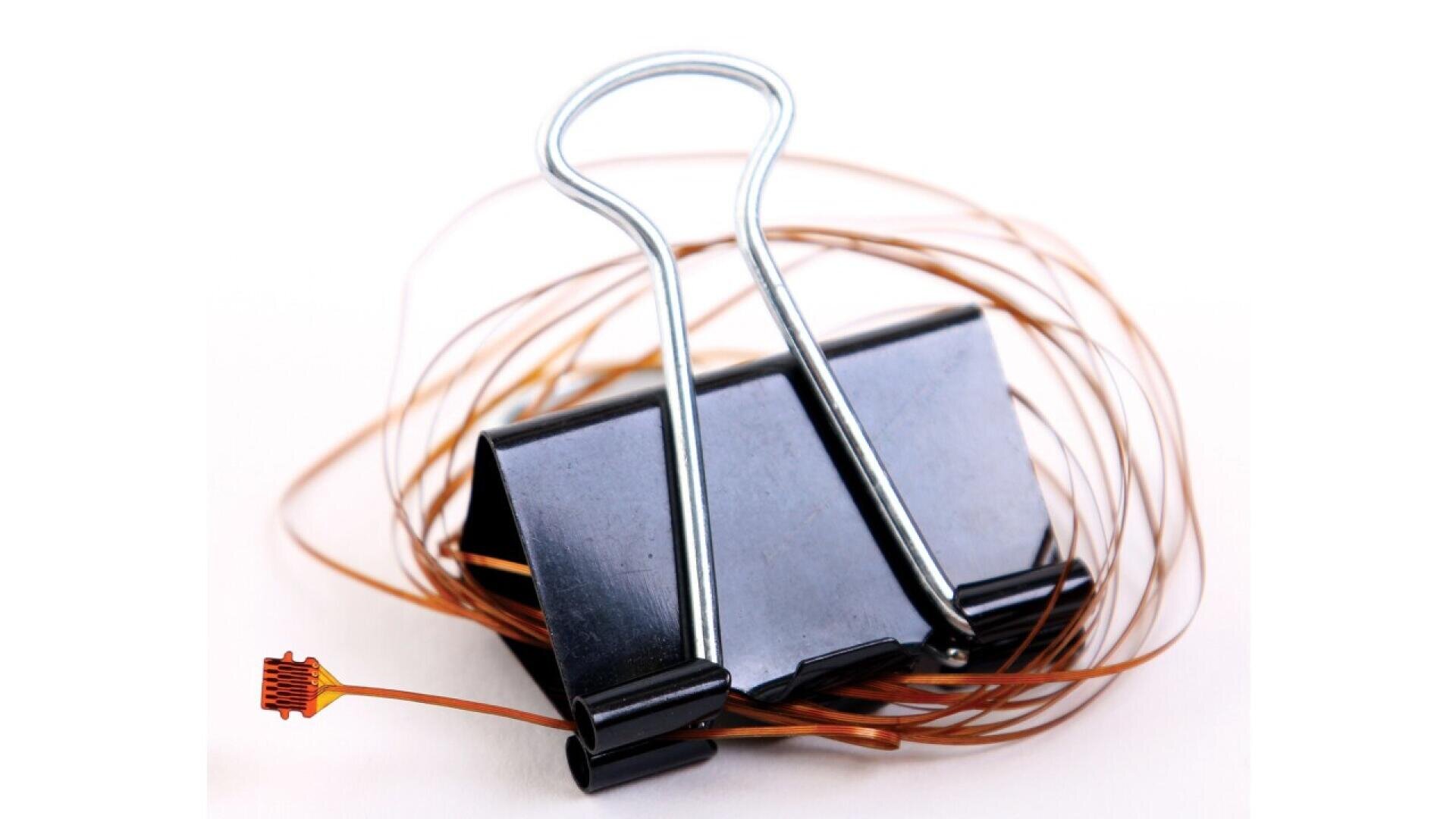 Eine schwarze Büroklammer hält vor einem weißen Hintergrund mehrere dünne Kupferdrähte, von denen einer in einem kleinen Sensor oder einer Sonde endet.
