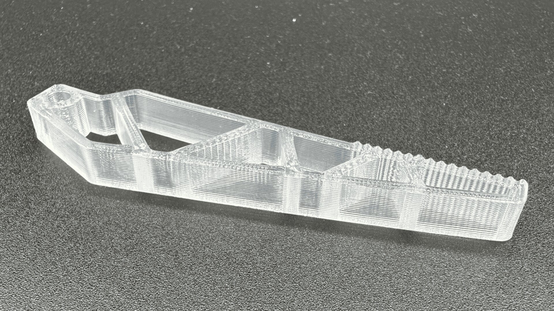Ein durchsichtiges, 3D-gedrucktes Kunststoffwerkzeug mit geometrischen Ausschnitten und geriffelten Kanten, platziert auf einer schwarzen strukturierten Oberfläche.