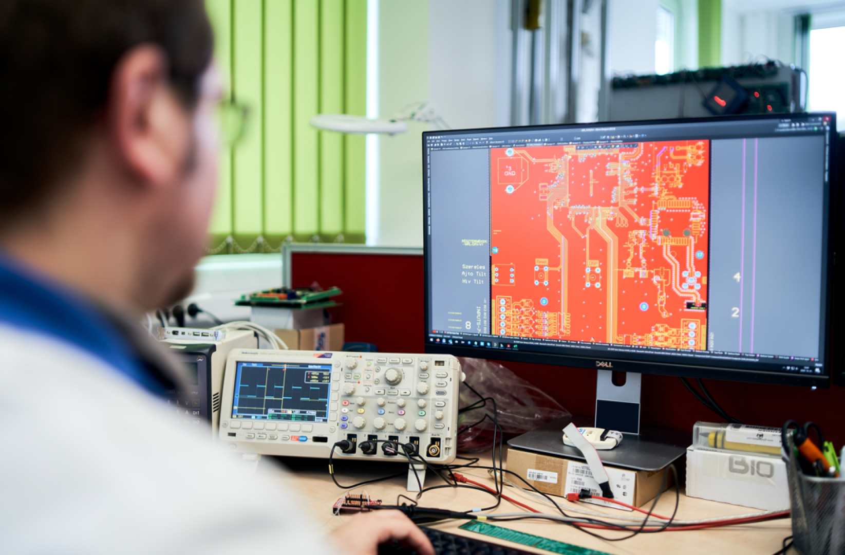 Eine Person arbeitet an einem Schreibtisch mit einem Computer, auf dem ein Entwurf einer elektronischen Leiterplatte angezeigt wird. Elektronische Prüfgeräte und Kabel liegen ebenfalls auf dem Schreibtisch. Im Hintergrund sind hellgrüne Jalousien zu sehen.