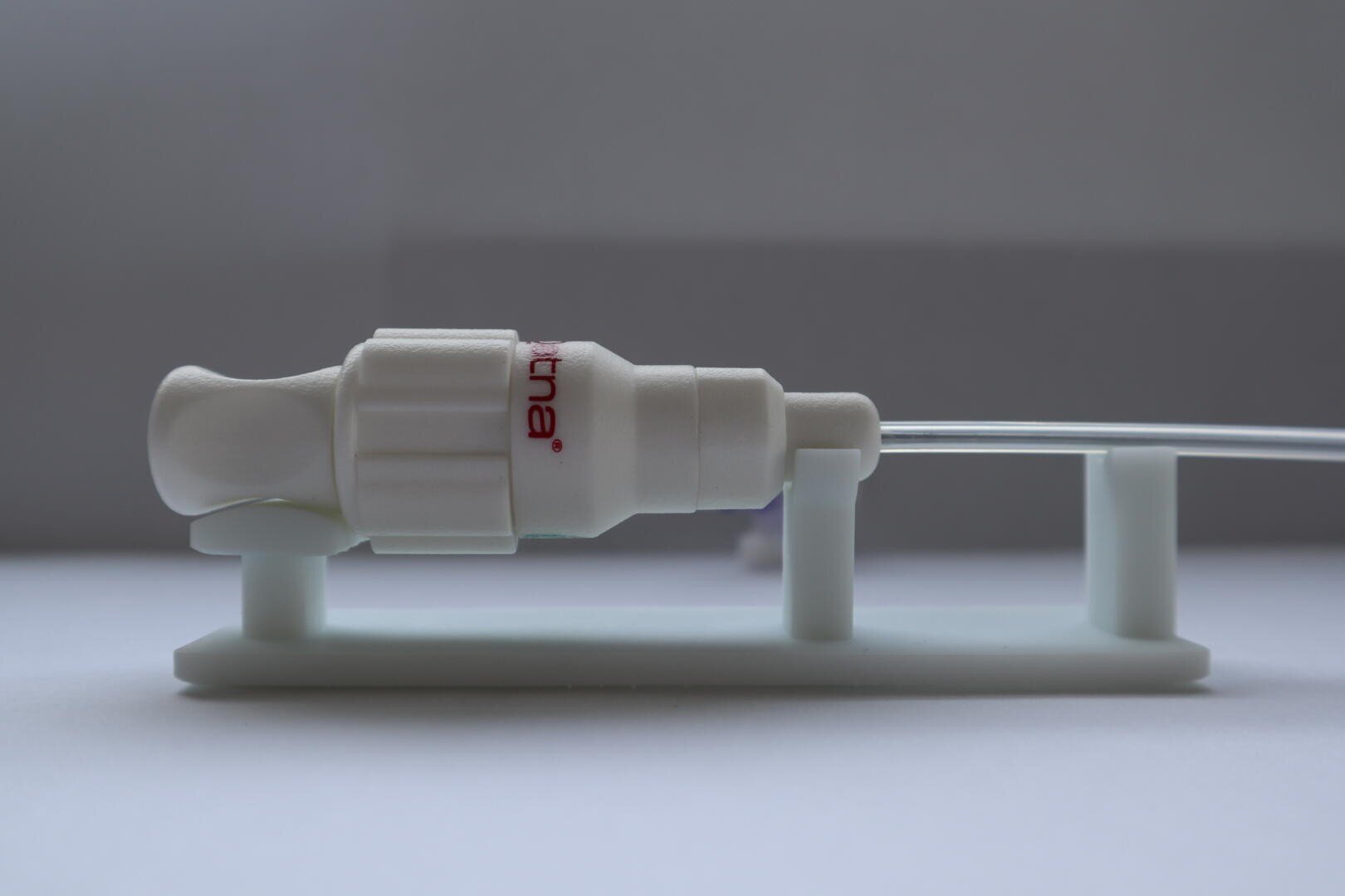 Nahaufnahme eines weißen medizinischen Konnektors, der an einem durchsichtigen Schlauch befestigt ist und auf einem weißen Kunststoffständer vor einem neutralen Hintergrund ruht.