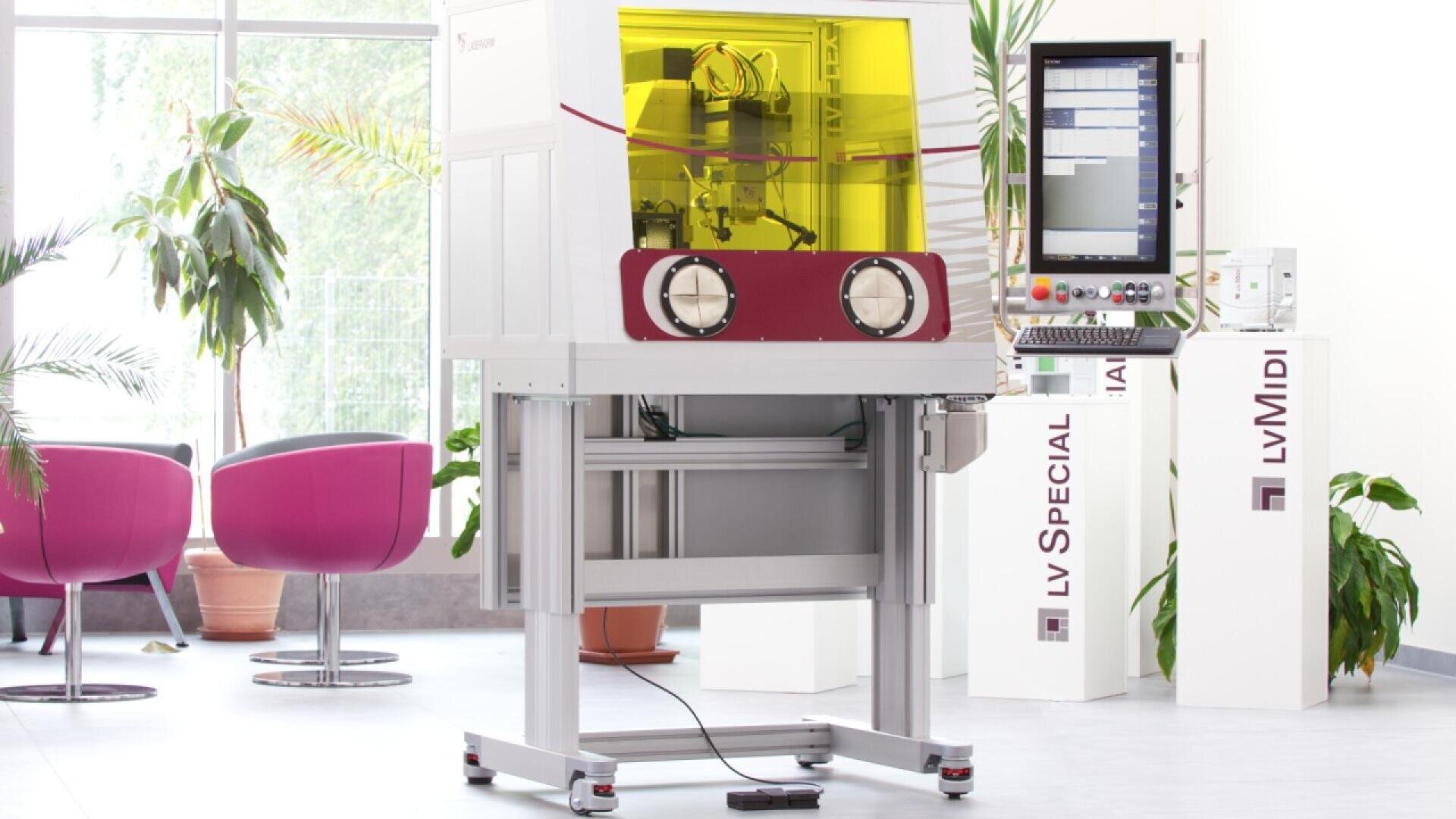 A modern laboratory machine with a yellow protective shield stands in a bright room with large windows, pink chairs, potted plants, and a control panel screen on a stand nearby.