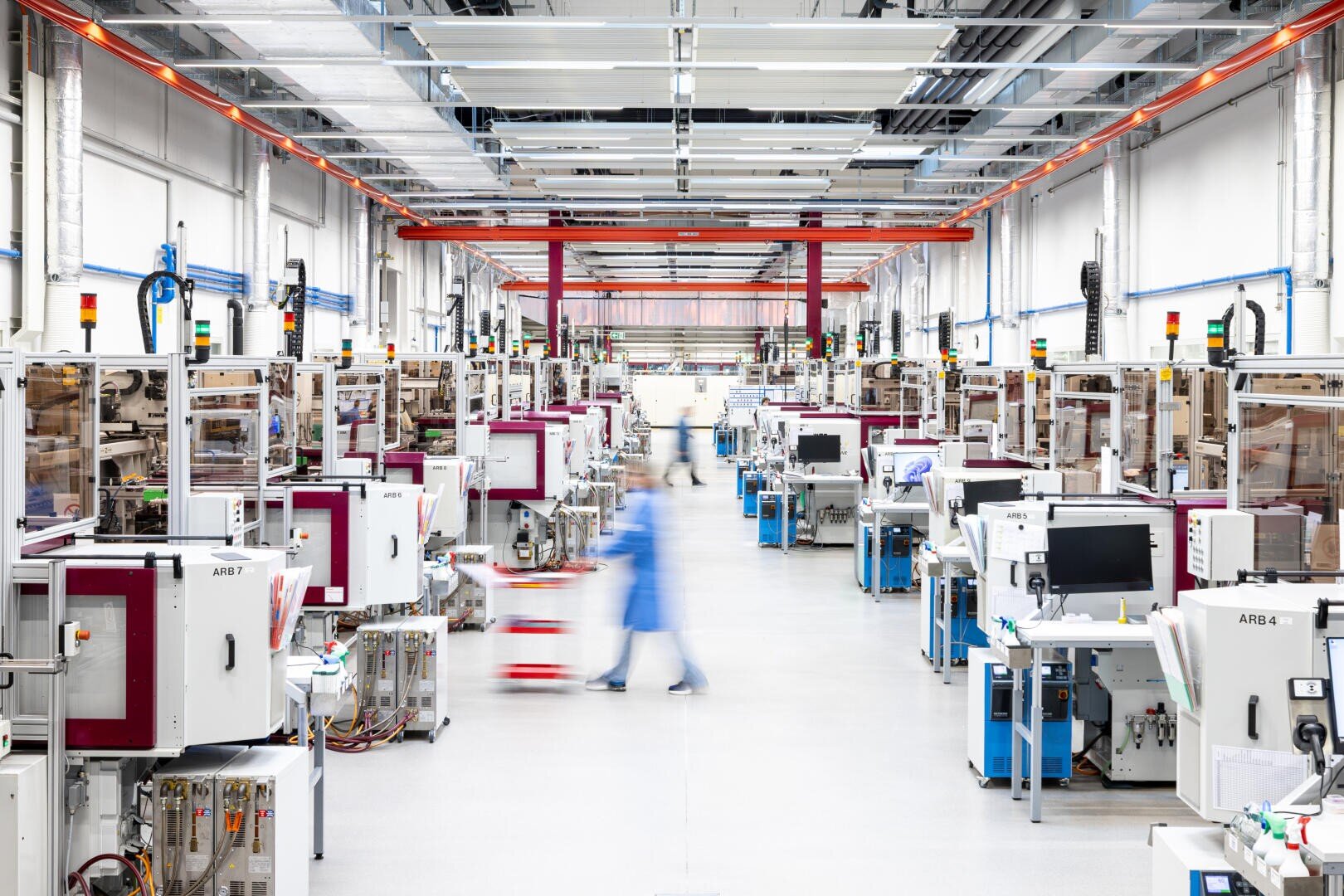 Wide view of a bright, modern factory floor with rows of industrial machines and equipment; a person in a blue coat walks down the center aisle, appearing blurred in motion.