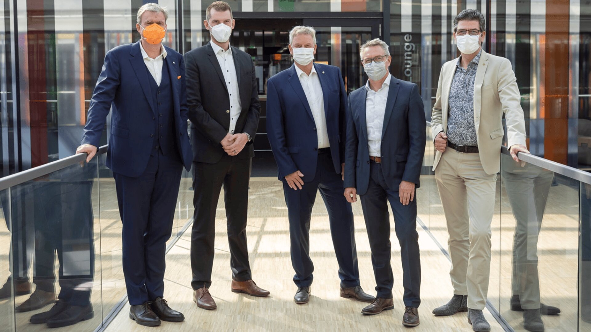 Fünf Männer in Anzügen und mit Gesichtsmasken stehen auf einem Glassteg in einem Innenraum und posieren für ein Gruppenfoto. Im Hintergrund ist moderne Architektur mit Glastüren und dem Wort "Lounge" zu sehen.