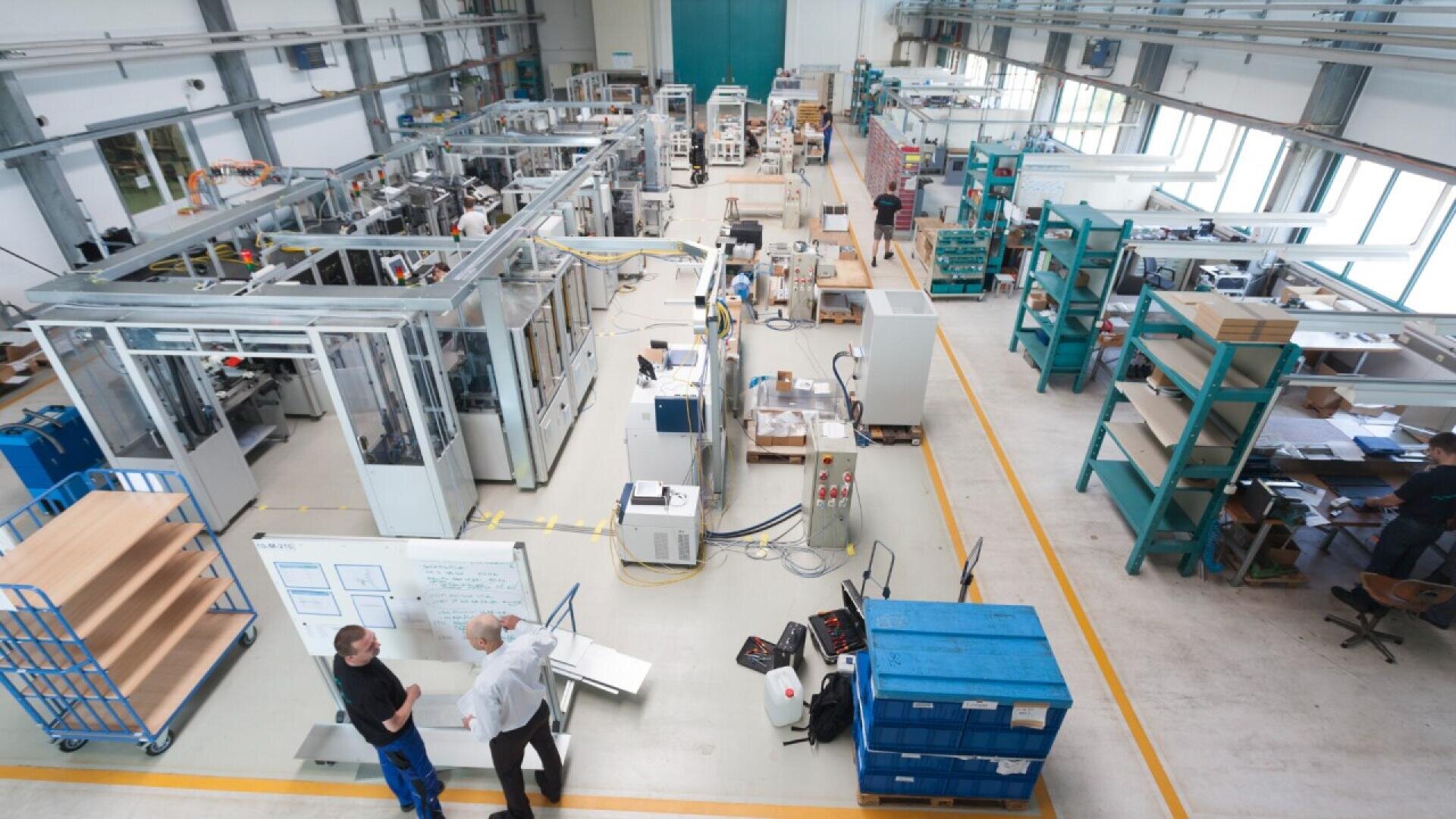 A wide view of a spacious, clean industrial workshop with various machinery, shelves, and equipment. Several people are working or conversing in different areas of the facility. Bright lighting and large windows are visible.