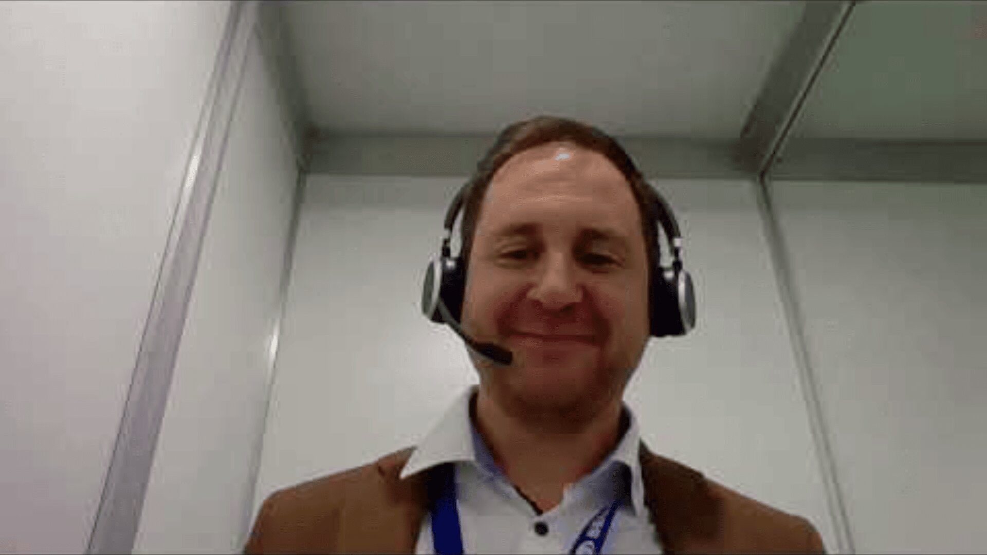 A man wearing a brown jacket, white shirt, and headset with a microphone is smiling while sitting in a modern, white-walled office or booth.