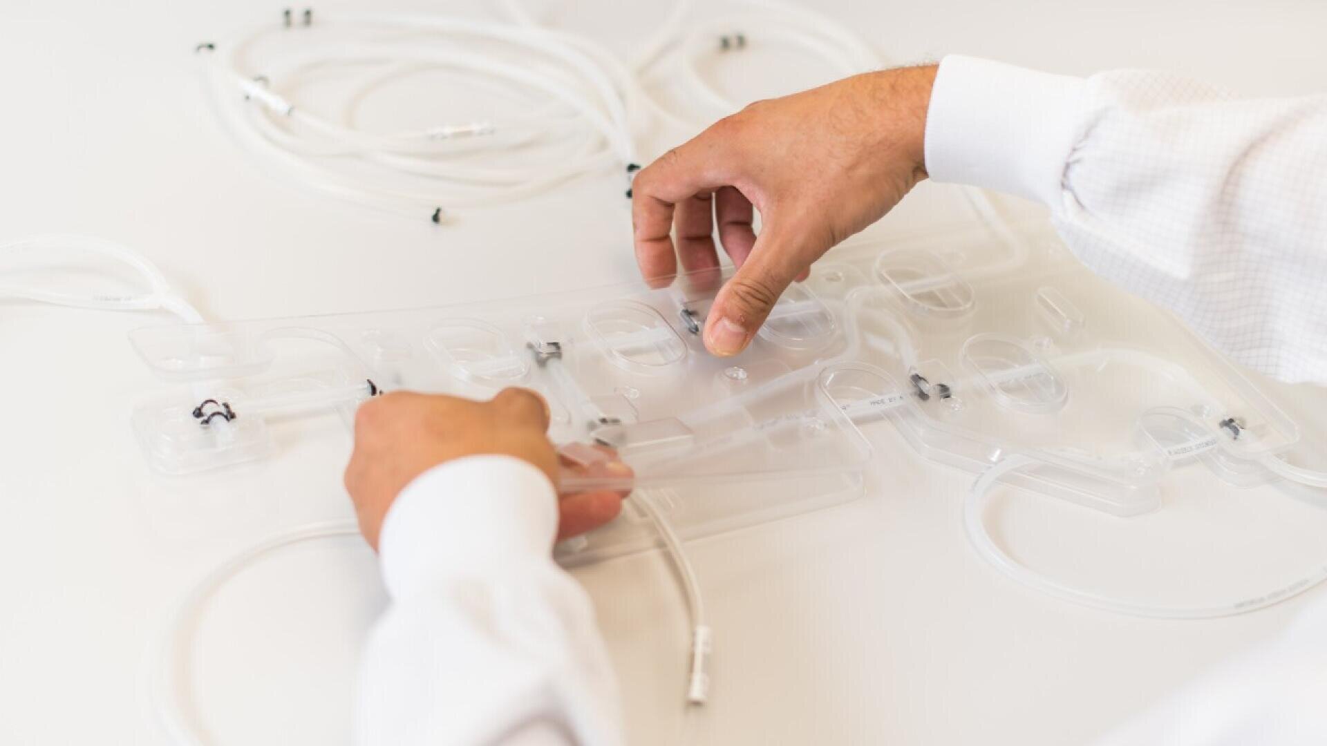 Die Hände einer Person richten medizinische Schläuche und Anschlüsse aus transparentem Kunststoff an einem transparenten Gerät aus, das auf einer weißen Oberfläche liegt. Die Person trägt ein weißes Langarmshirt.