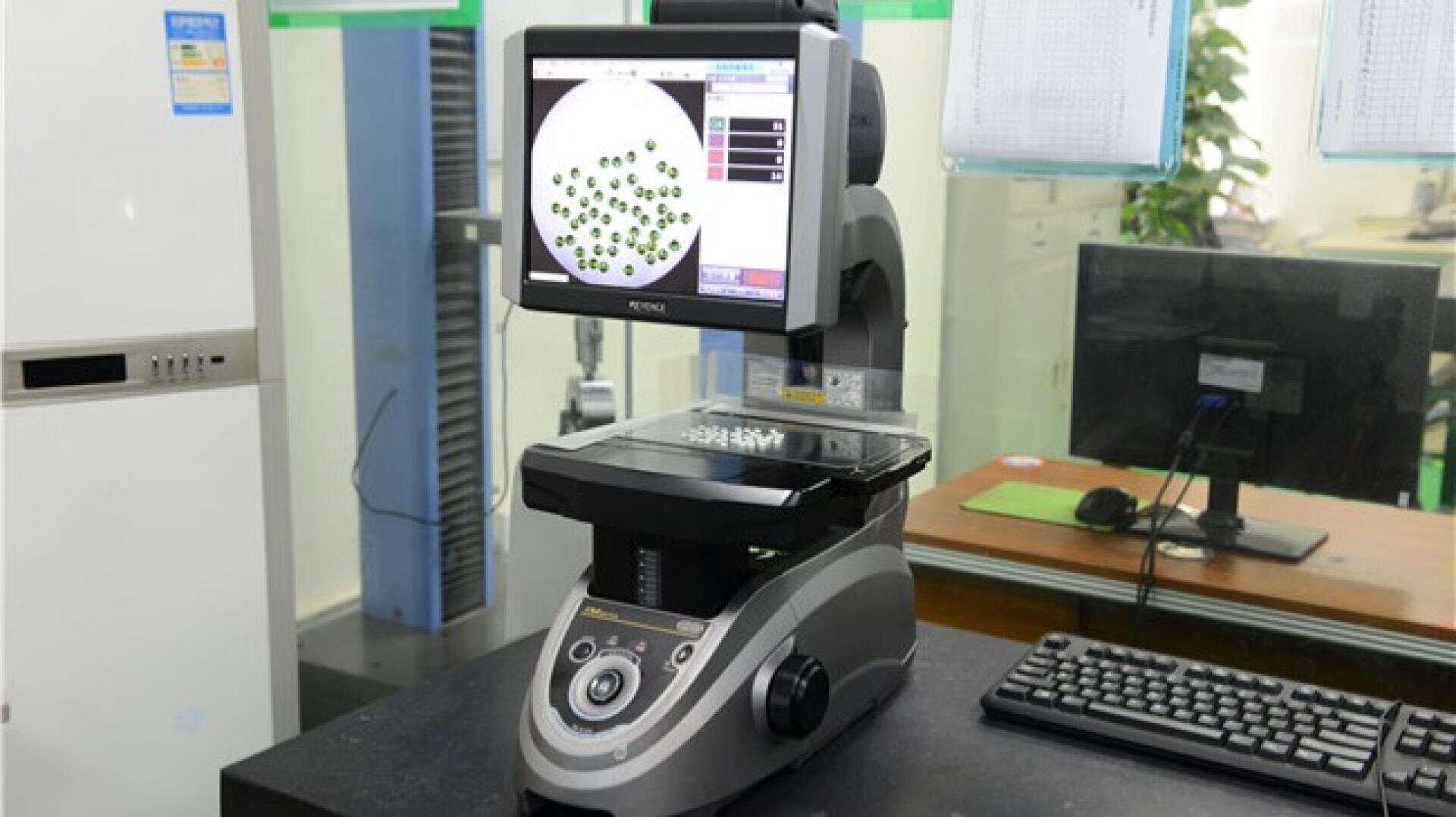 A digital measuring microscope displays magnified circular objects on its screen in a laboratory setting, with a keyboard nearby and charts pinned on a glass wall in the background.