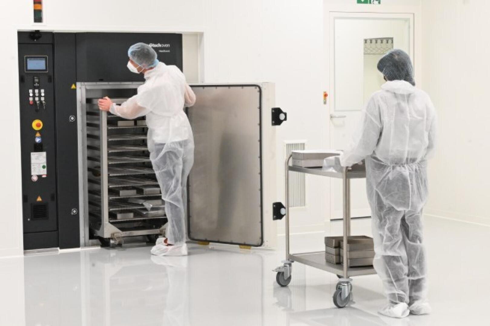 Two people in full protective clothing work in a cleanroom; one opens a large industrial machine with trays inside, while the other stands by a trolley with metal trays.