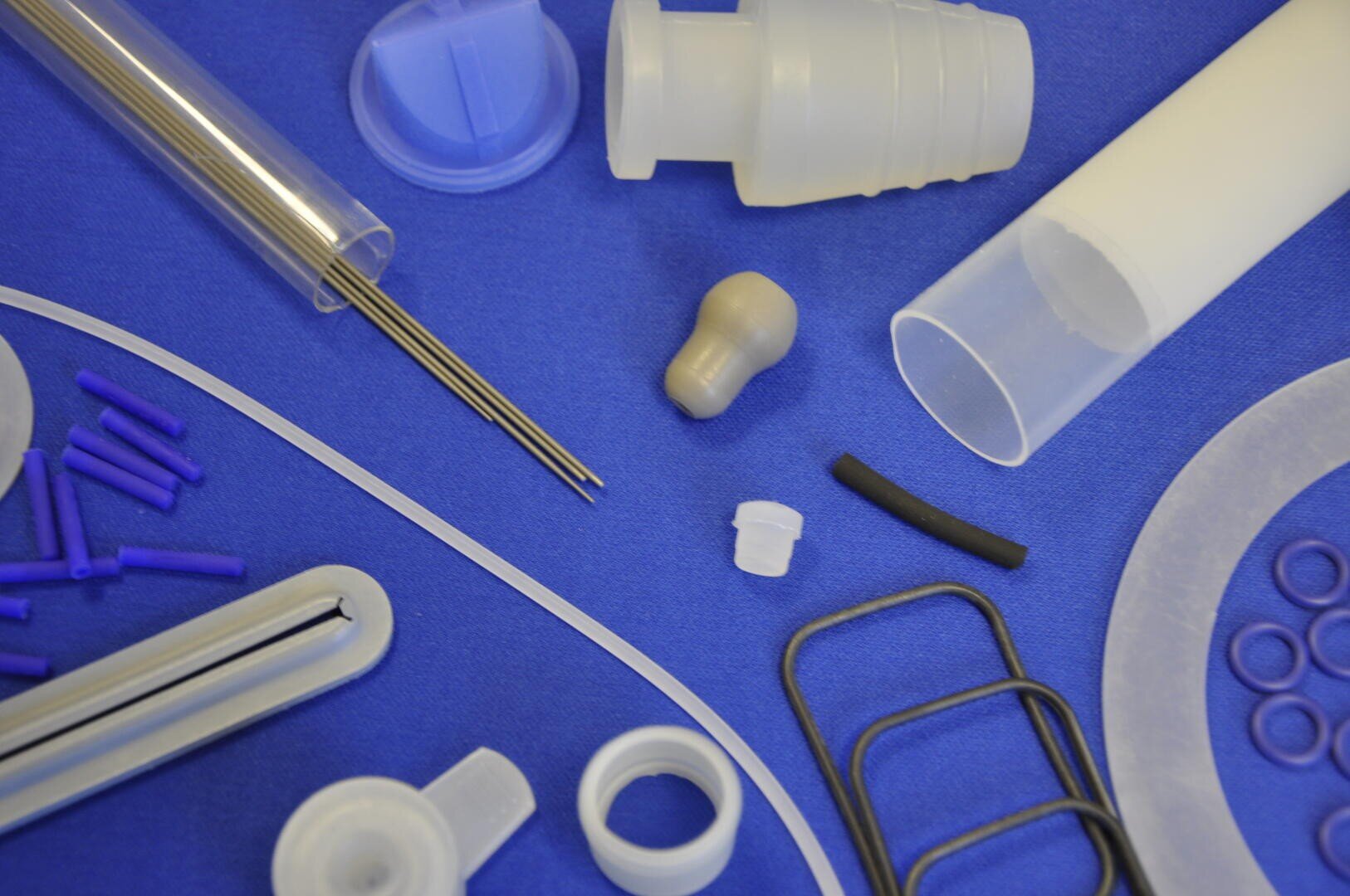 A variety of small plastic and metal medical components, rods, tubes, rings, and seals are arranged on a blue surface. The items are assorted in shape, size, and color, mainly white, clear, purple, and black.