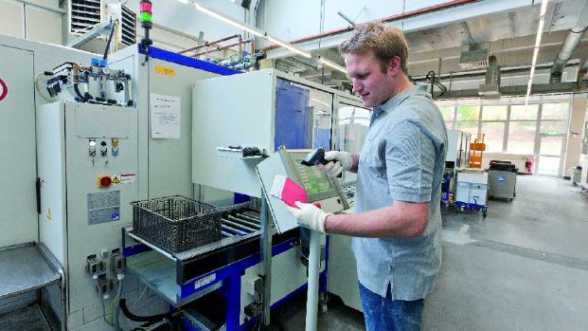 Ein Mann mit Handschuhen bedient ein Bedienfeld in einer hell erleuchteten Industrieanlage und steht neben einer großen Maschine mit Metallteilen und Geräten im Hintergrund.