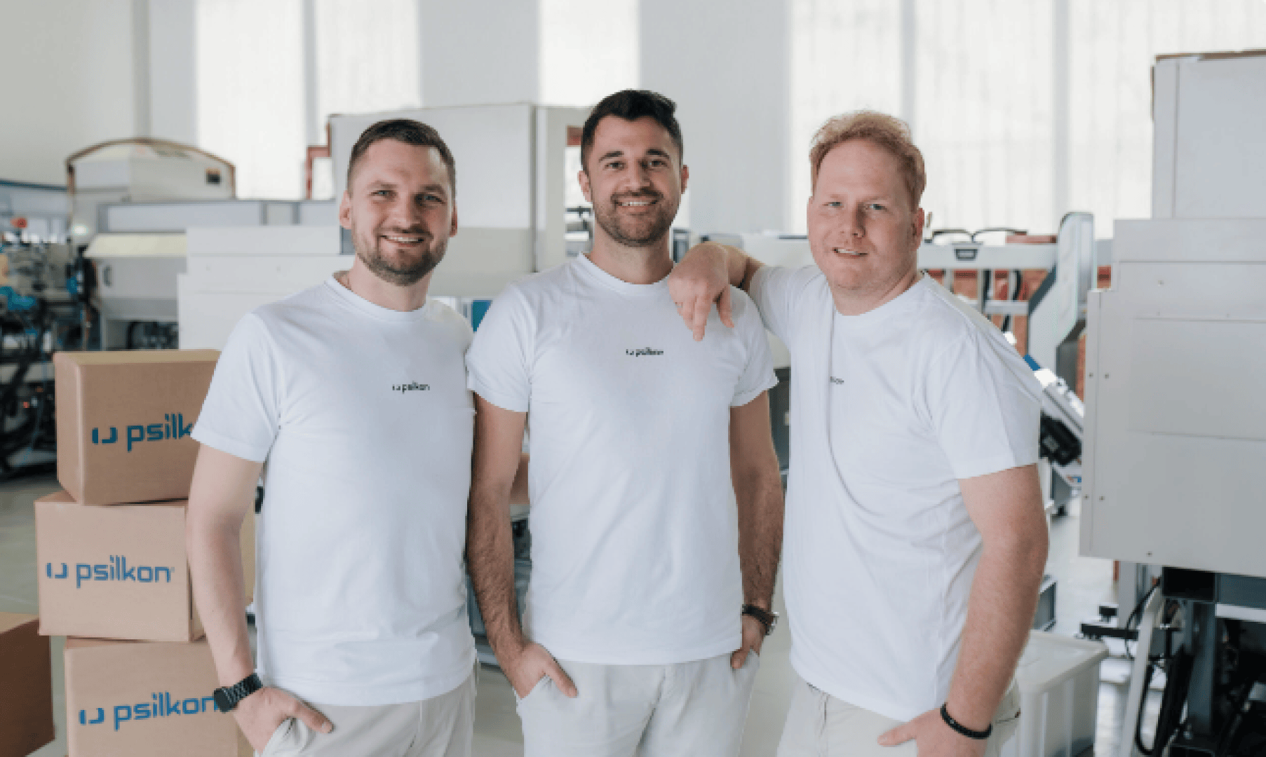Three men wearing matching white t-shirts stand together smiling in a bright industrial or office space with boxes and equipment in the background. One man has his arm around another’s shoulder.
