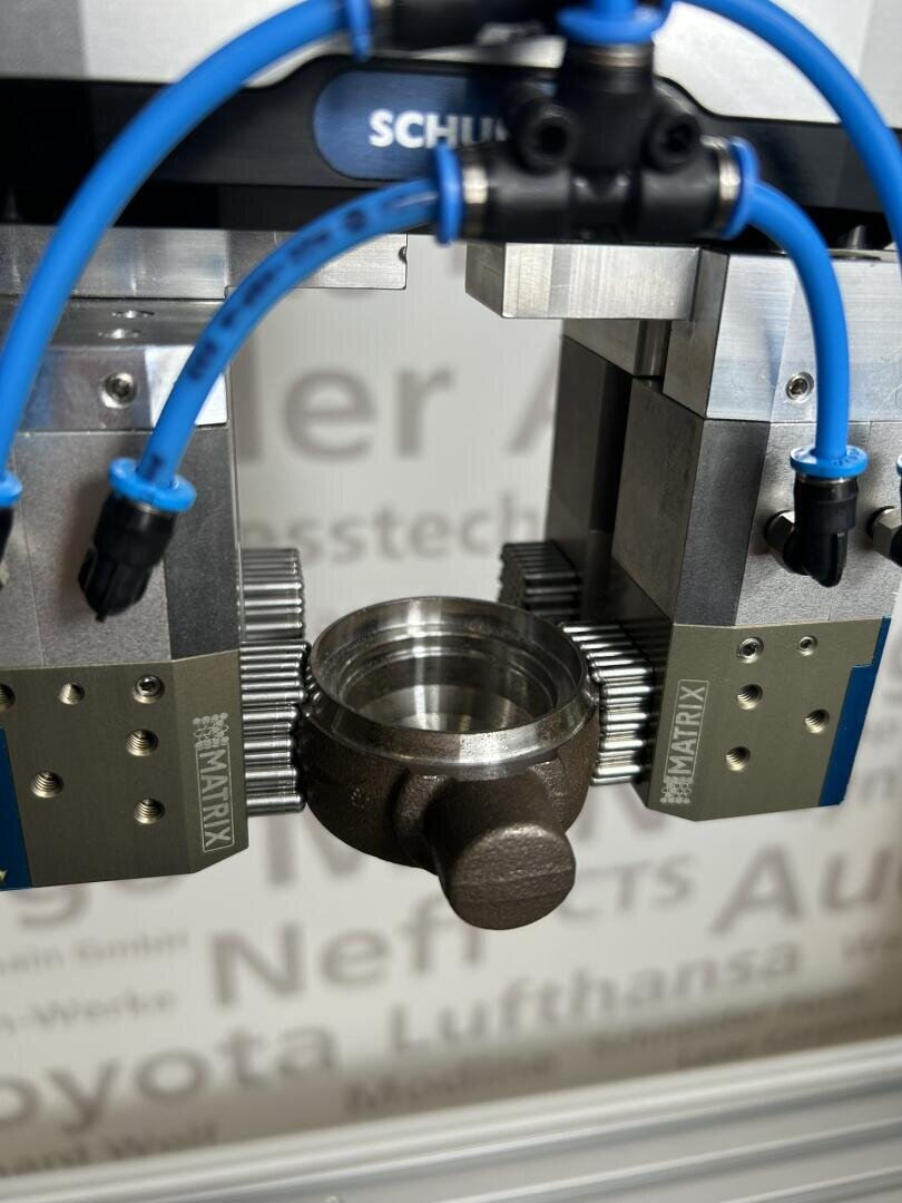 Ein Robotergreifer mit blauen Kabeln hält ein Metallteil in einer Fertigungs- oder Industrieumgebung. Der Hintergrund ist unscharf mit teilweise sichtbarem Text an einer Wand.