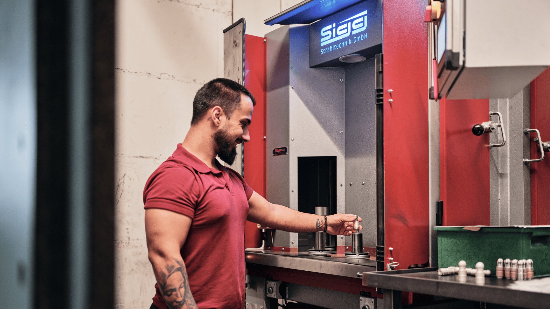 Ein Mann im roten Poloshirt bedient eine rot-graue Industriemaschine. Im Inneren hält er ein kleines zylindrisches Teil. Auf einem blau beleuchteten Bildschirm darüber zeigt die Maschine das Wort SIGS an.