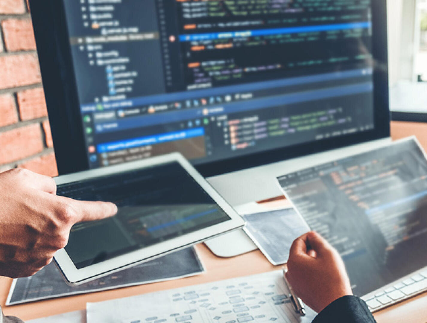 Zwei Personen arbeiten an einem Schreibtisch mit einem großen Computerbildschirm, auf dem Code angezeigt wird. Eine Person hält ein Tablet in der Hand, die andere arbeitet mit einem Laptop. Beide prüfen Programmier- und technische Dokumente.