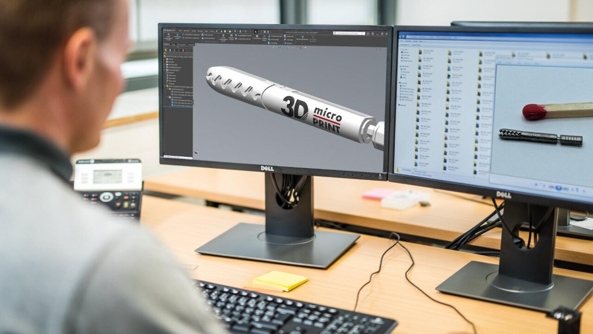 A person sits at a desk with dual monitors; one screen shows a 3D CAD model labeled 3D micro PRINT, and the other displays a photo comparison of a small metal part next to a matchstick.