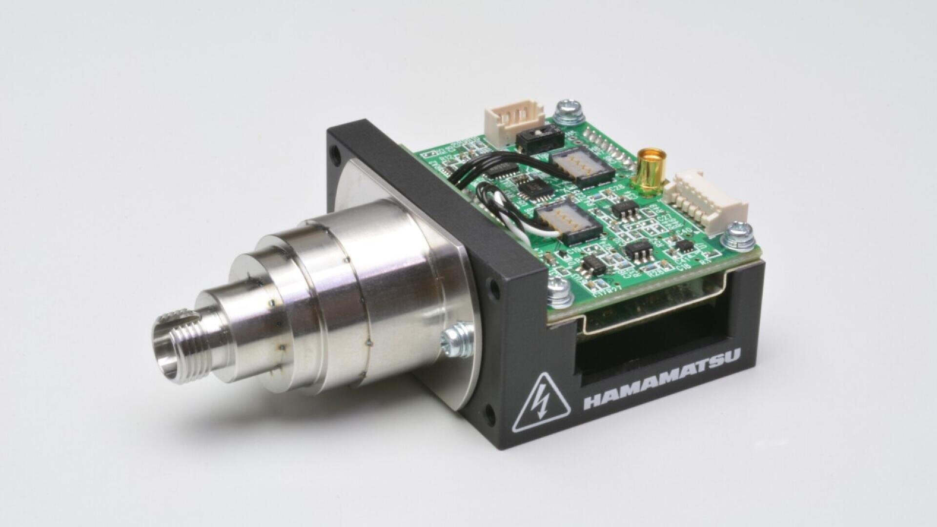 A compact electronic device with a metal cylindrical lens and exposed circuit board, labeled Hamamatsu, placed on a light gray surface.