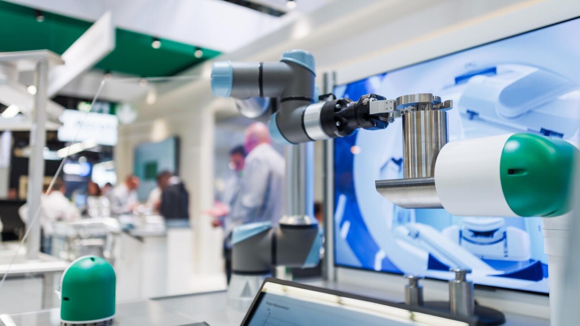 A close-up of a robotic arm with a blue and gray design in a modern tech exhibition, with people and large digital screens blurred in the background.