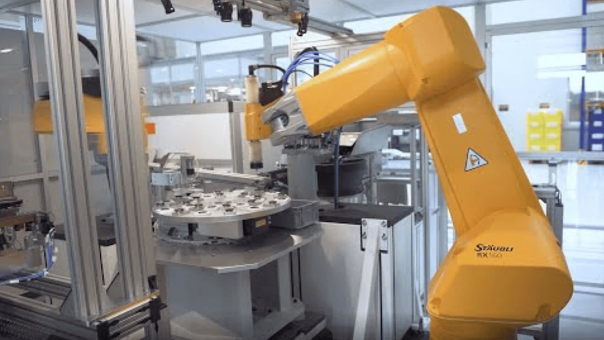 A yellow industrial robotic arm operates in a modern automated factory, handling components on a rotary table surrounded by glass panels and metal structures.