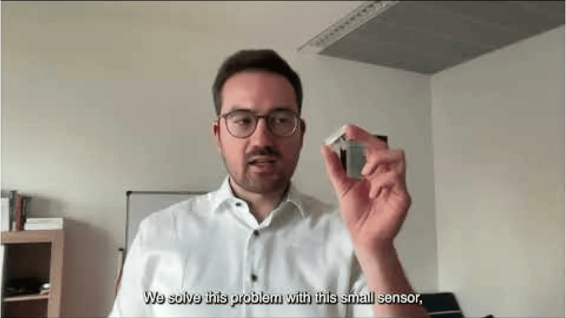 Ein Mann mit Brille und weißem Hemd hält ein kleines Sensorgerät in der Hand. Der Untertitel lautet: „Wir lösen dieses Problem mit diesem kleinen Sensor.“ Er steht in einem modernen Büro.