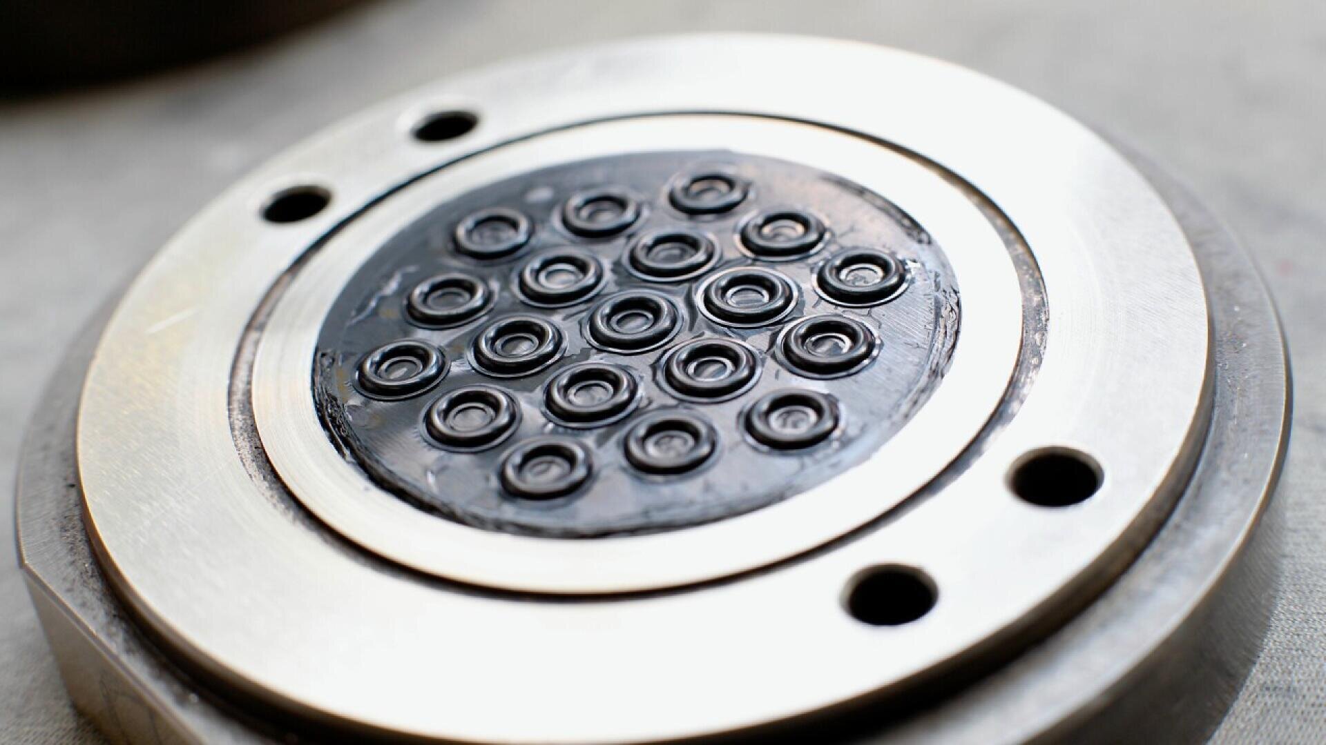 Close-up of a metallic circular device with multiple small circular openings arranged in a pattern in the center, surrounded by a shiny metal ring and two larger holes on the outer edge.