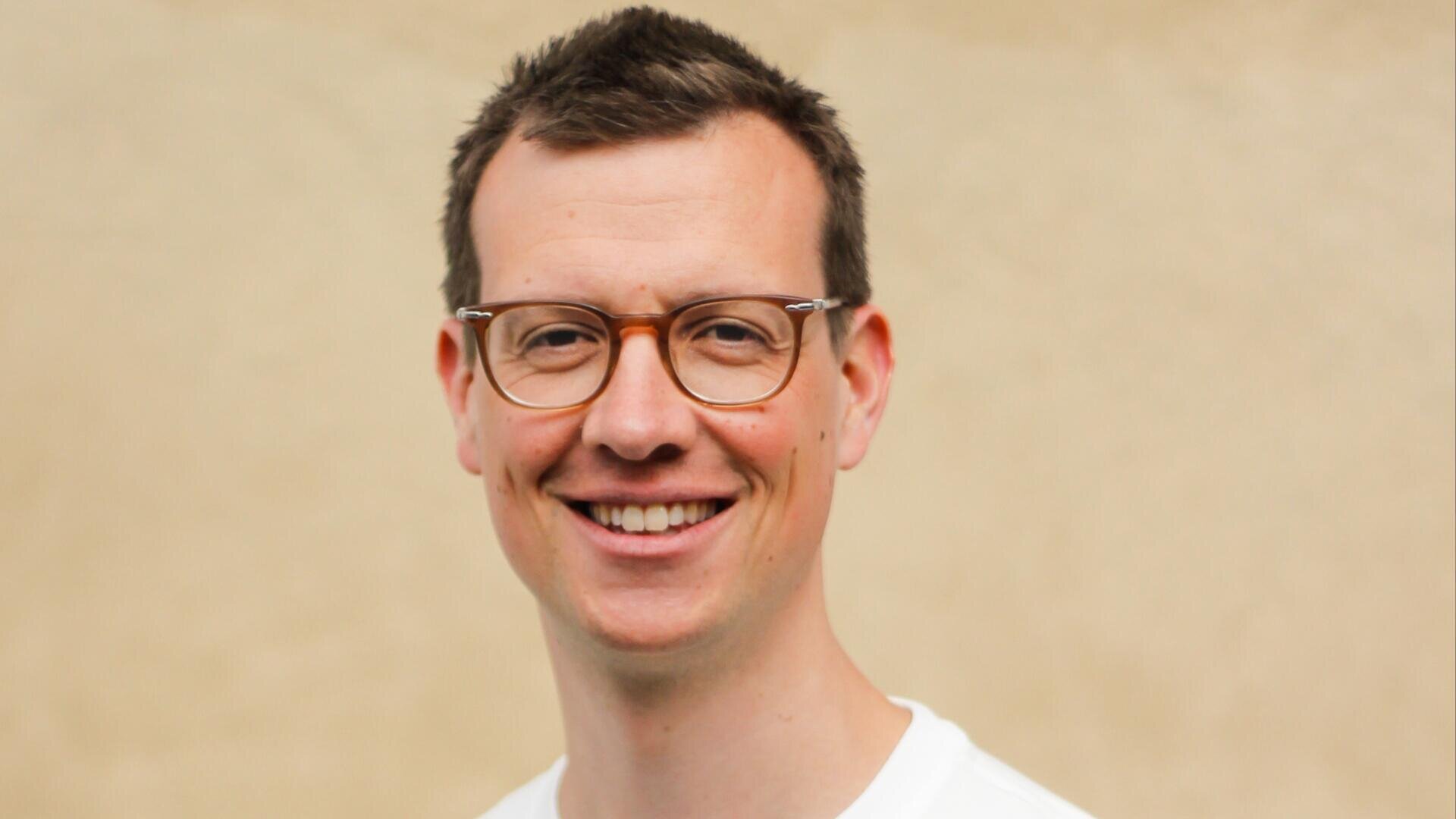 Ein Mann mit kurzen braunen Haaren und Brille lächelt in die Kamera. Er trägt ein weißes Hemd und steht vor einem schlichten beigen Hintergrund.