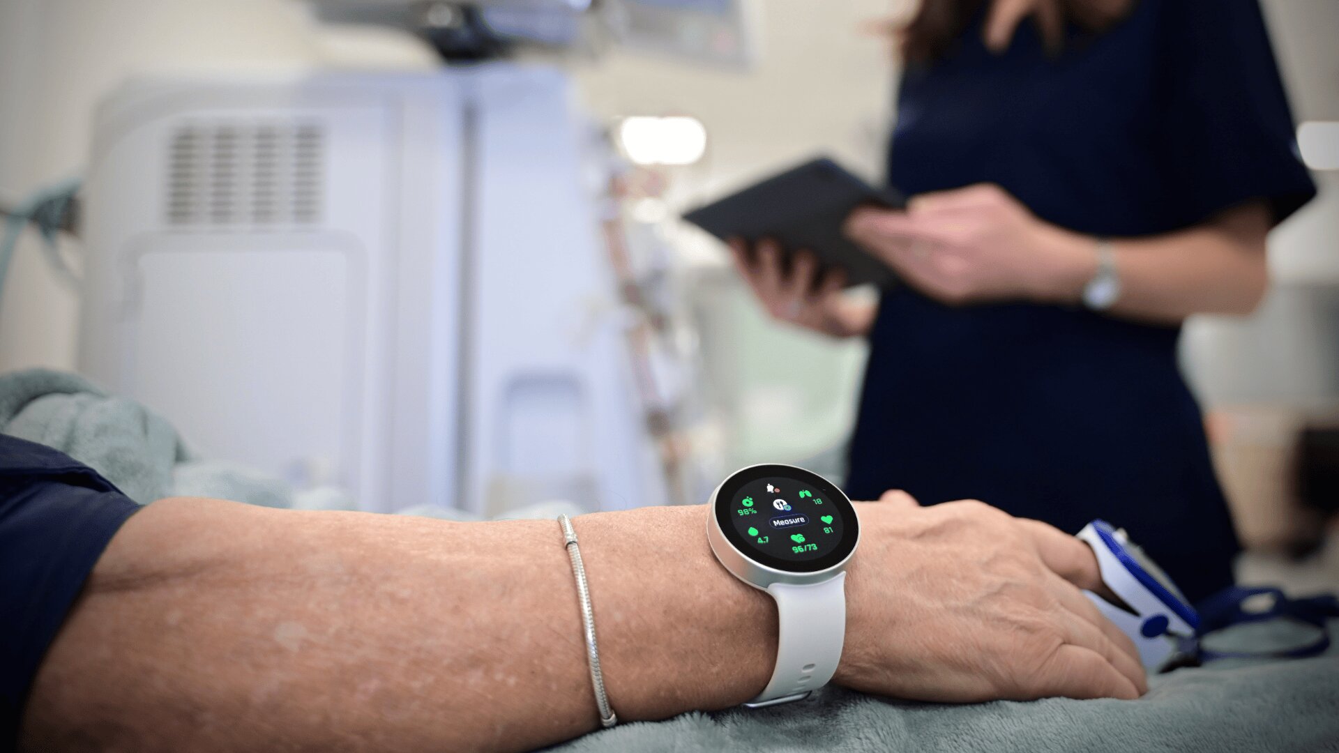 A person’s hand wearing a white smartwatch rests on a blanket in a medical setting, with medical equipment nearby and a healthcare worker holding a tablet in the blurred background.