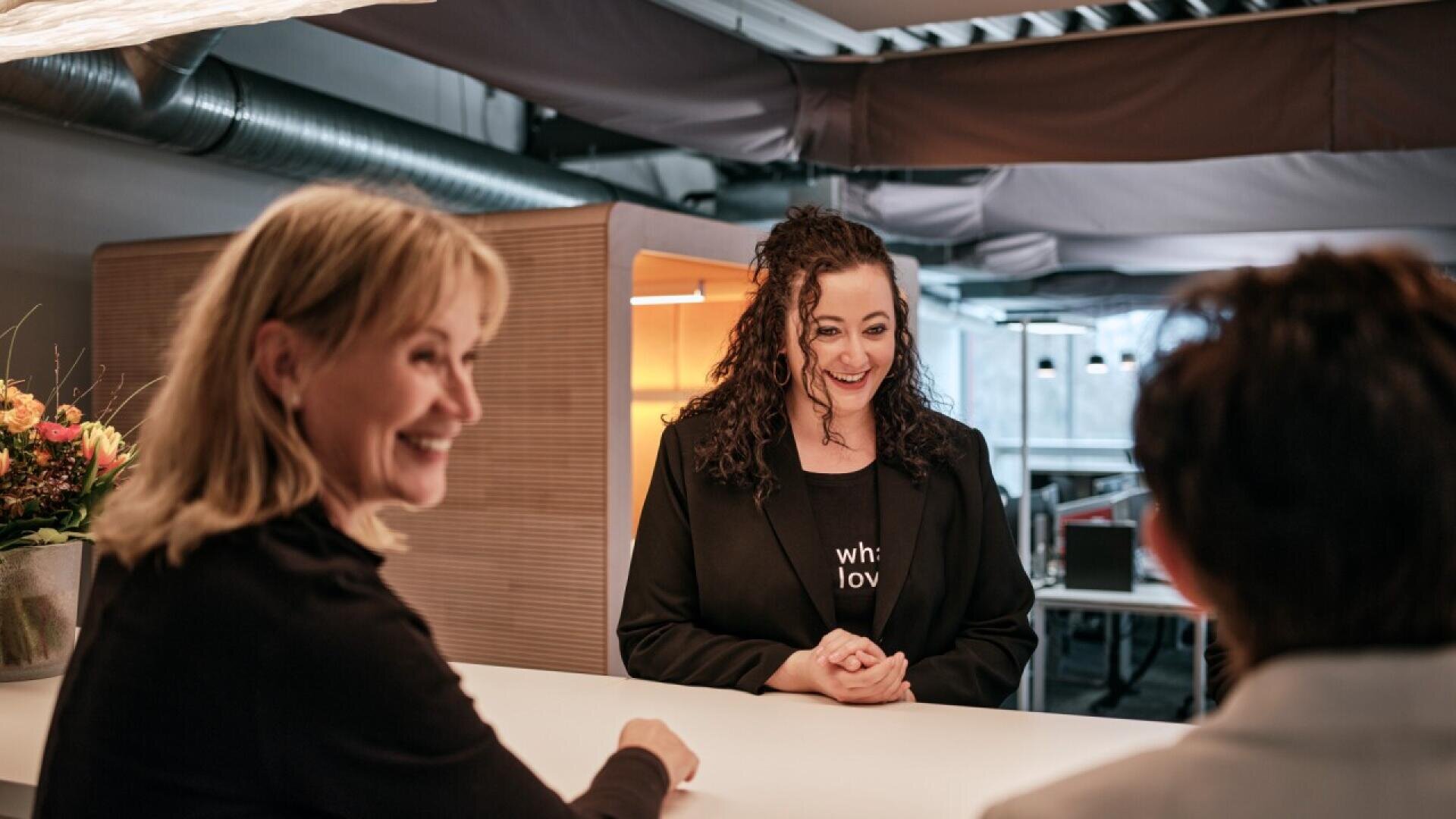 Three women are having a friendly conversation at a modern reception desk; one woman stands smiling behind the counter, while two others sit across from her, facing away from the camera.