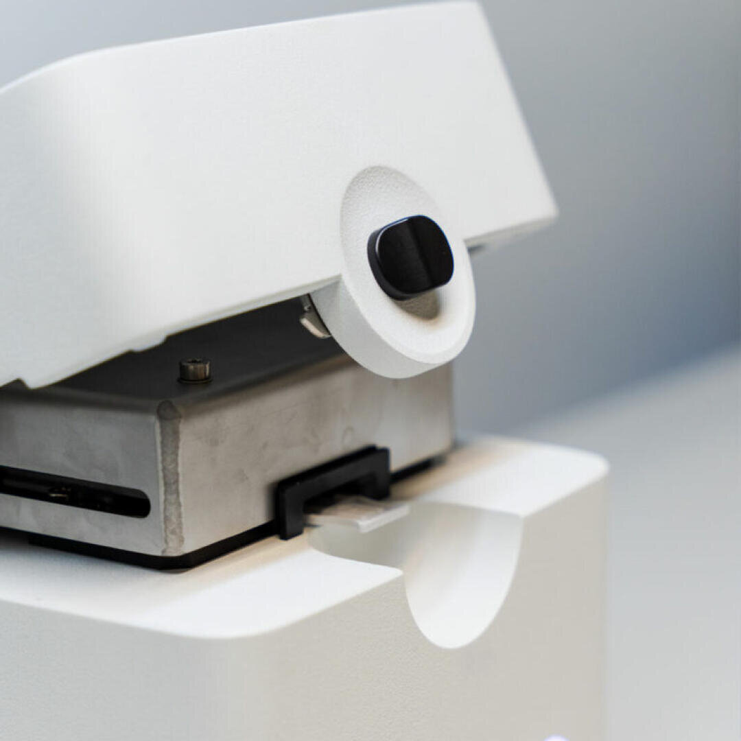 Close-up of a white laboratory instrument with a black button and metal component, likely a microtome used for cutting thin sections of samples for microscopic examination. The background is out of focus.