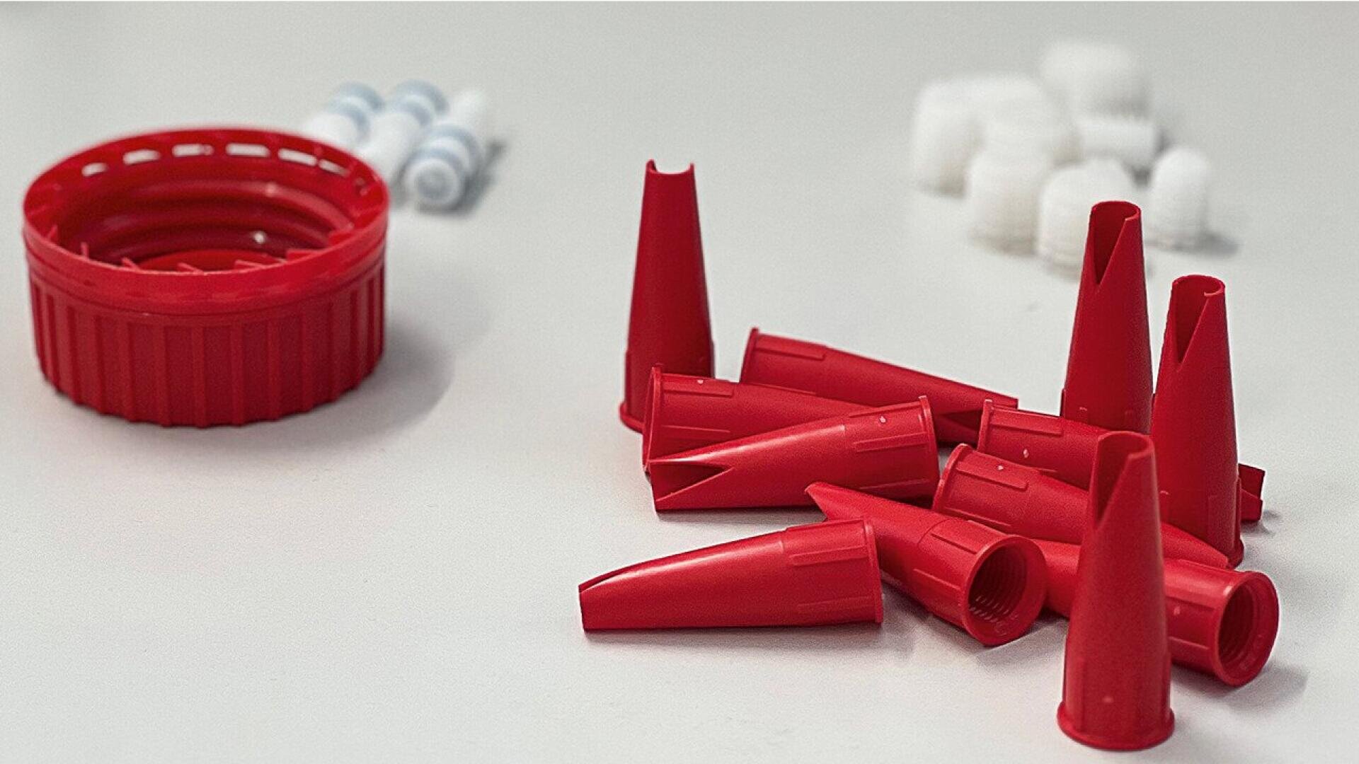 Several red plastic caps and a large red lid are scattered on a white surface, with small white and blue caps blurred in the background.