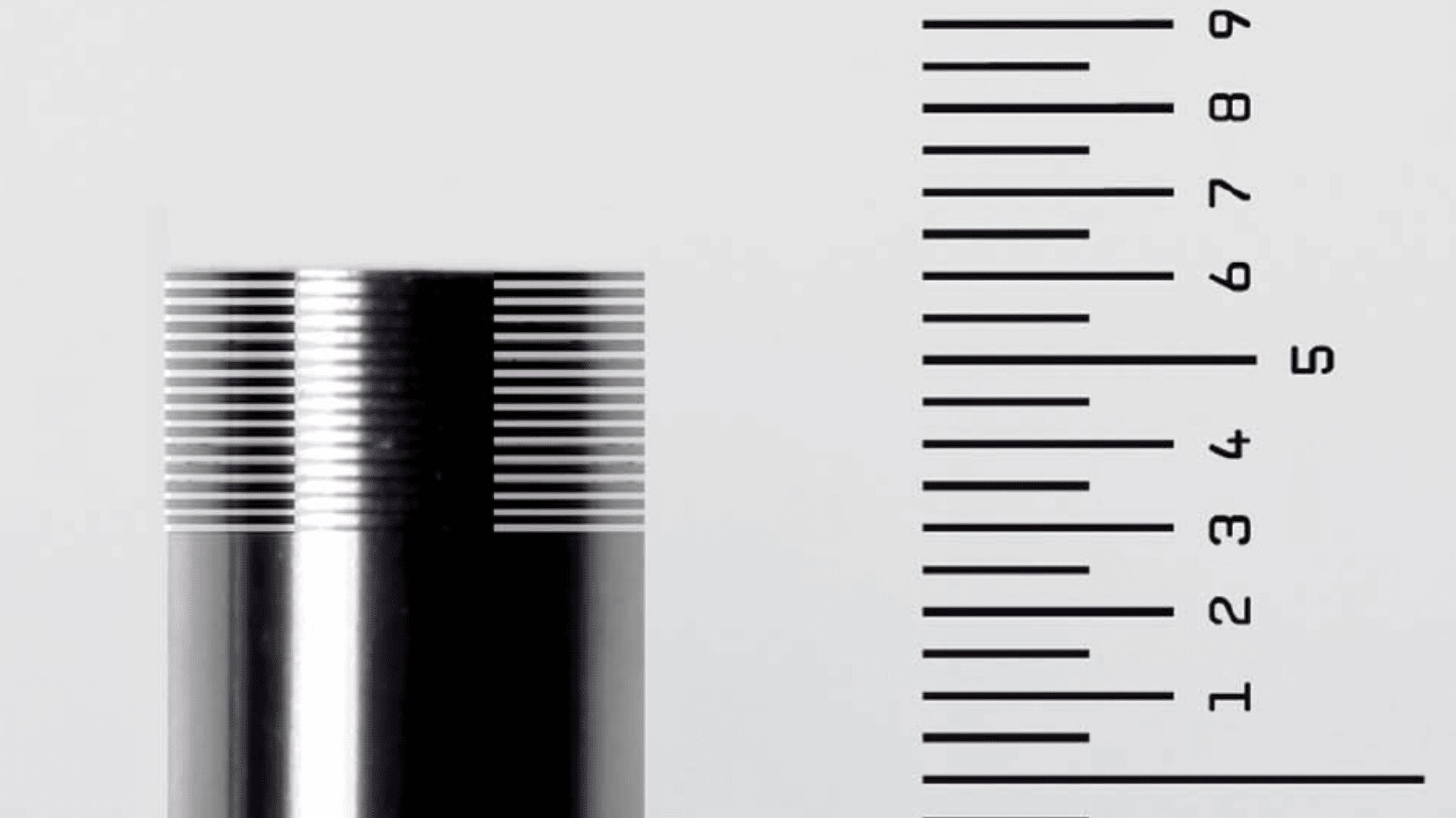 Eine Nahaufnahme eines Metallzylinders mit feinen horizontalen Rillen neben einem vertikalen Lineal mit den Zahlen 1 bis 9 und gleichmäßig verteilten schwarzen Linien auf hellem Hintergrund.