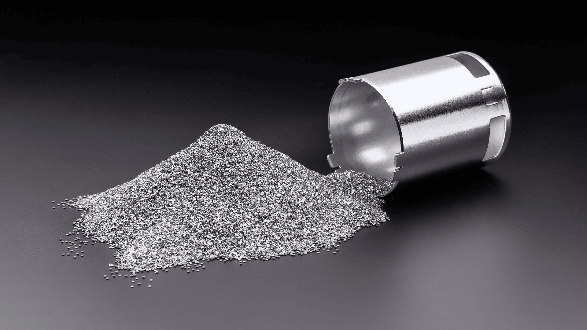 A small metal container tipped over on its side, spilling a pile of shiny metallic granules onto a smooth, black surface.