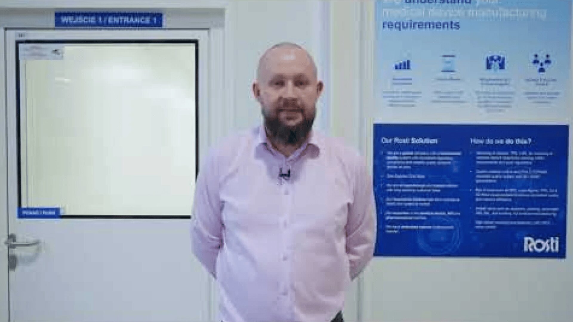 A man with a beard, wearing a light pink shirt, stands indoors near a white door labeled Entrance 1. Behind him is a blue and white informational poster on the wall.