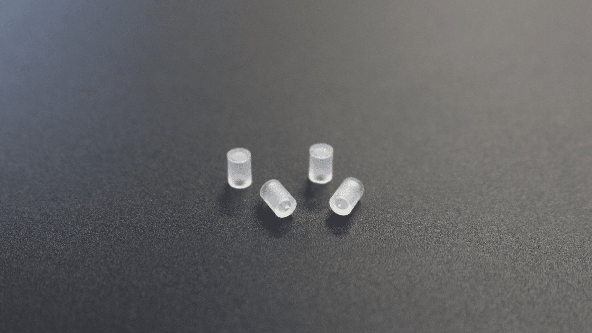 Vier kleine, durchscheinende, zylindrische Kunststoffkappen sind auf einer glatten, dunklen Oberfläche angeordnet.