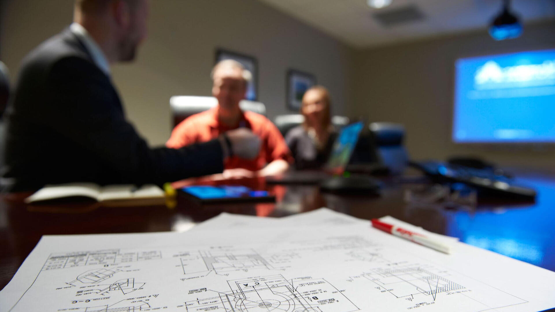 Three people sit around a conference table with technical drawings and a red pen in focus. A notebook and laptop are also on the table, and a presentation is projected on a screen in the background.