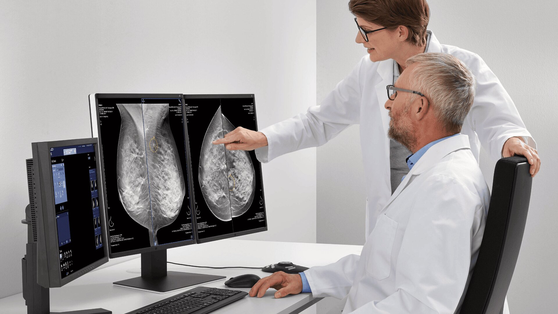 Two doctors in white lab coats review breast X-ray images on dual computer monitors. One doctor is seated, while the other stands, pointing at details on one of the screens in a medical office setting.