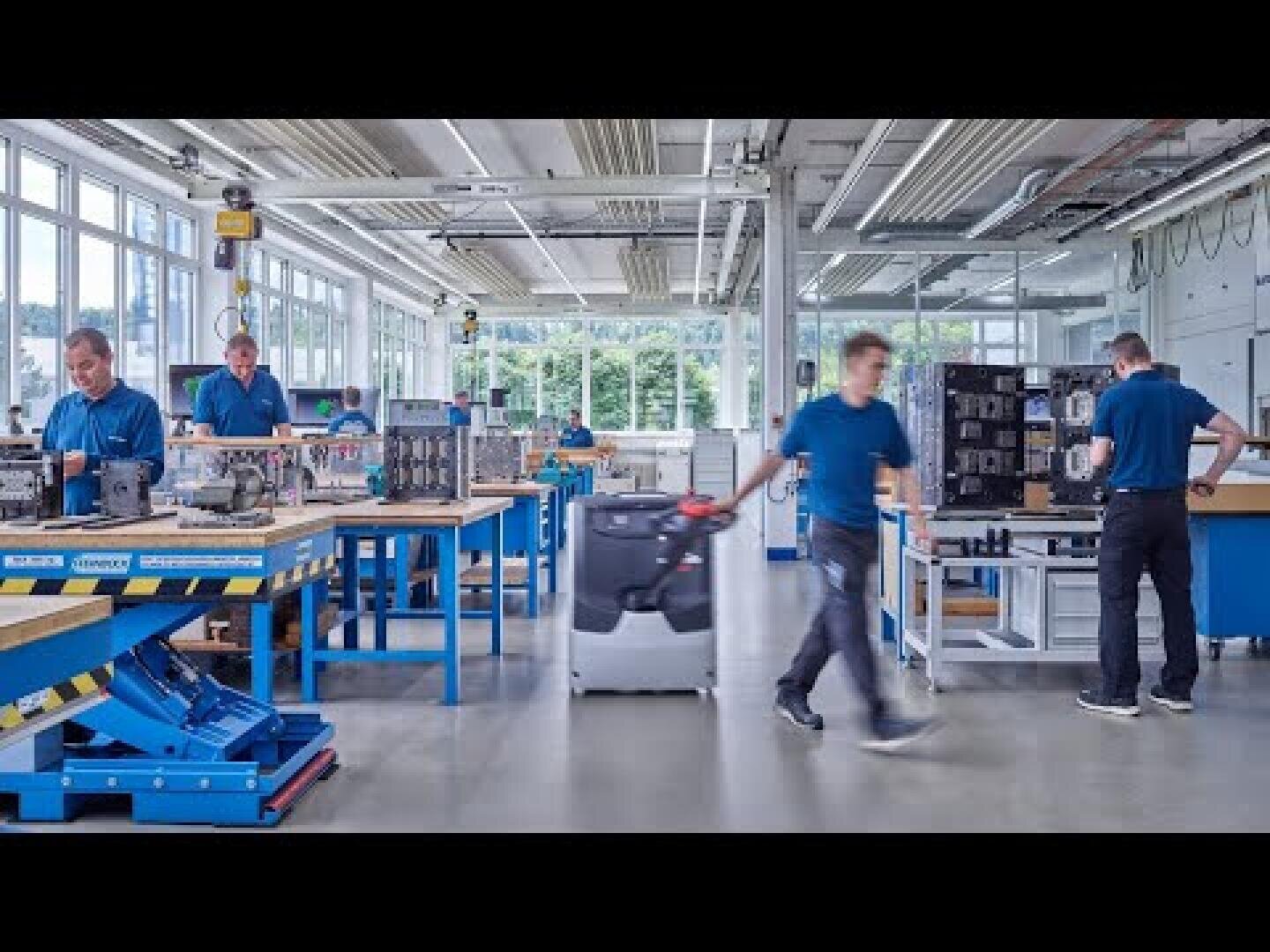 Eine helle, moderne Fabrikhalle mit mehreren Personen in blauen Hemden, die an Bänken arbeiten, Maschinen bedienen und Geräte bewegen. Große Fenster lassen natürliches Licht herein, und verschiedene Werkzeuge und Komponenten sind zu sehen.