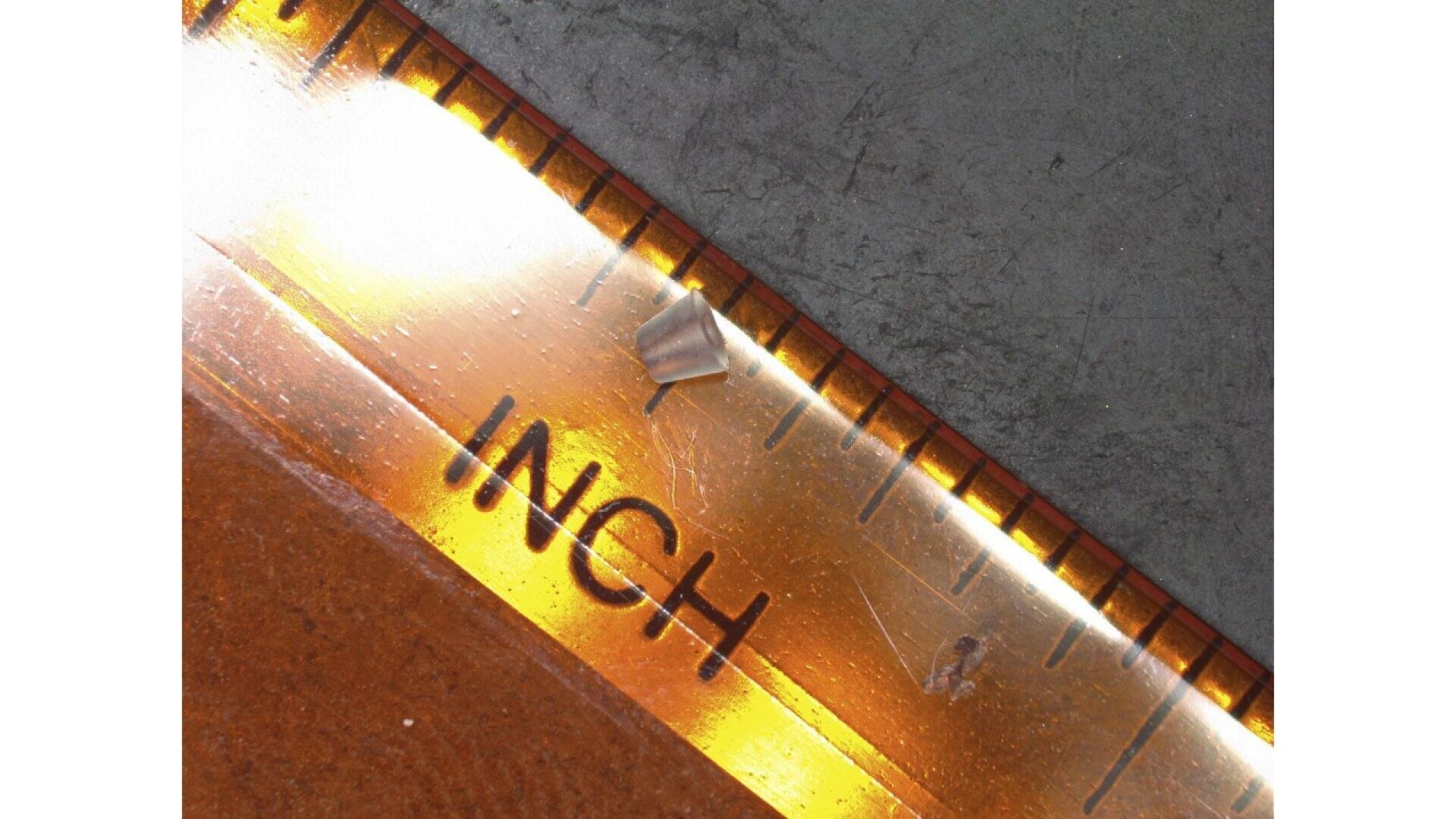 A small metallic splinter rests on the 1-inch mark of a transparent ruler, with the word INCH clearly visible and a textured gray surface in the background.