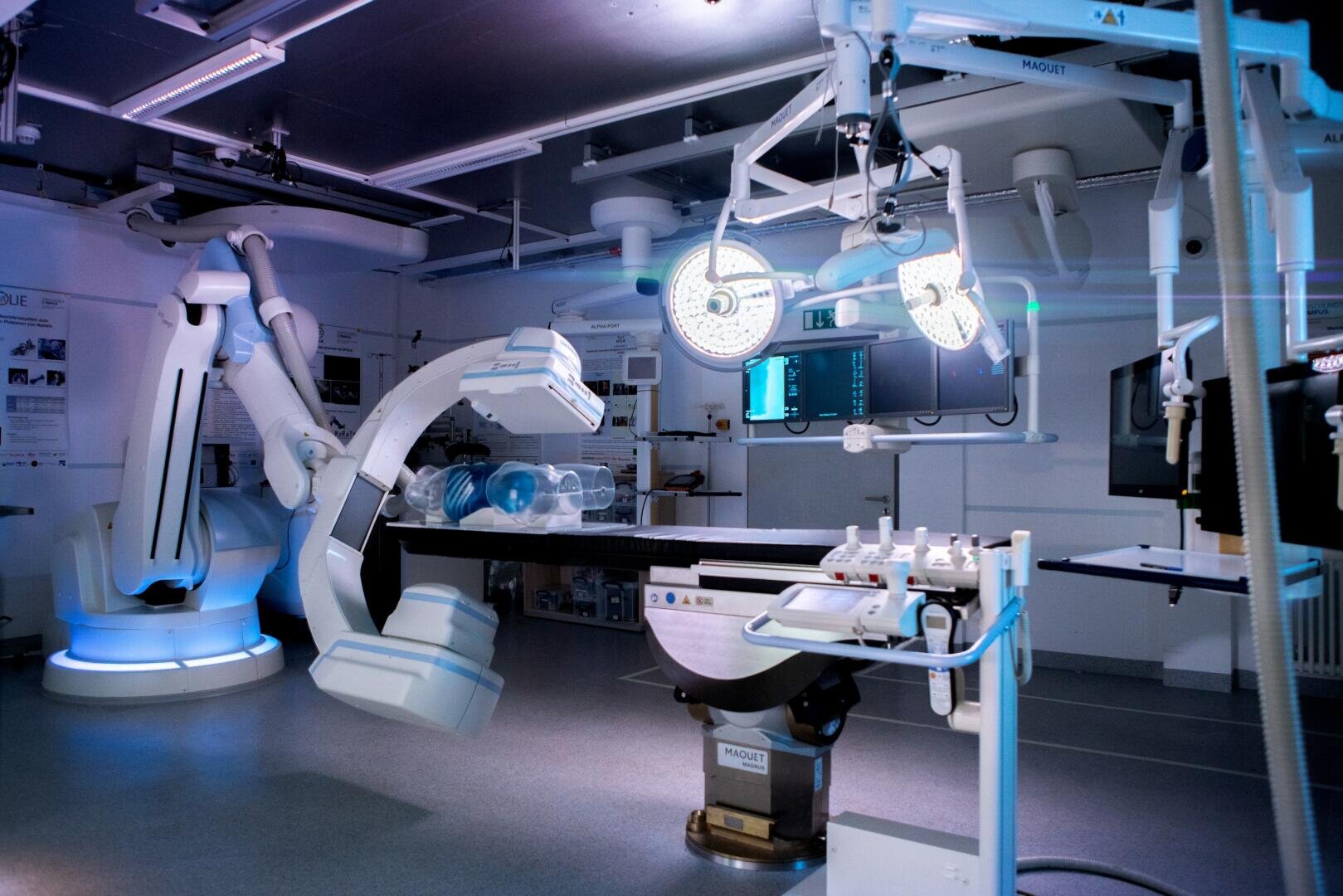 A modern hospital operating theater with advanced medical equipment, robotic surgical arms, bright ceiling lights and an empty operating table, all illuminated by cool blue and white light.