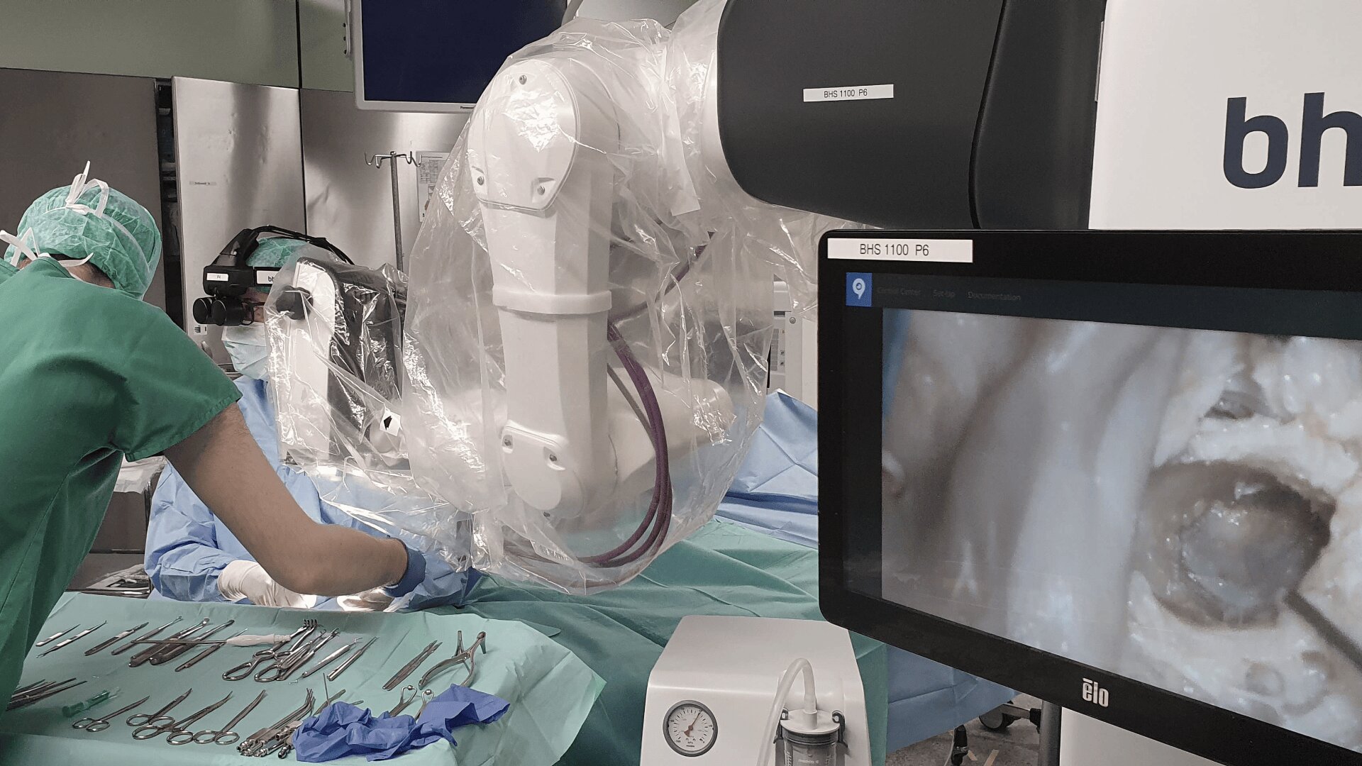 Ein Chirurg in grünem Kittel operiert neben einem mit Kunststoff verkleideten Operationsroboter, auf dem chirurgische Instrumente ausgelegt sind und ein Monitor ein medizinisches Bild in Großaufnahme zeigt.