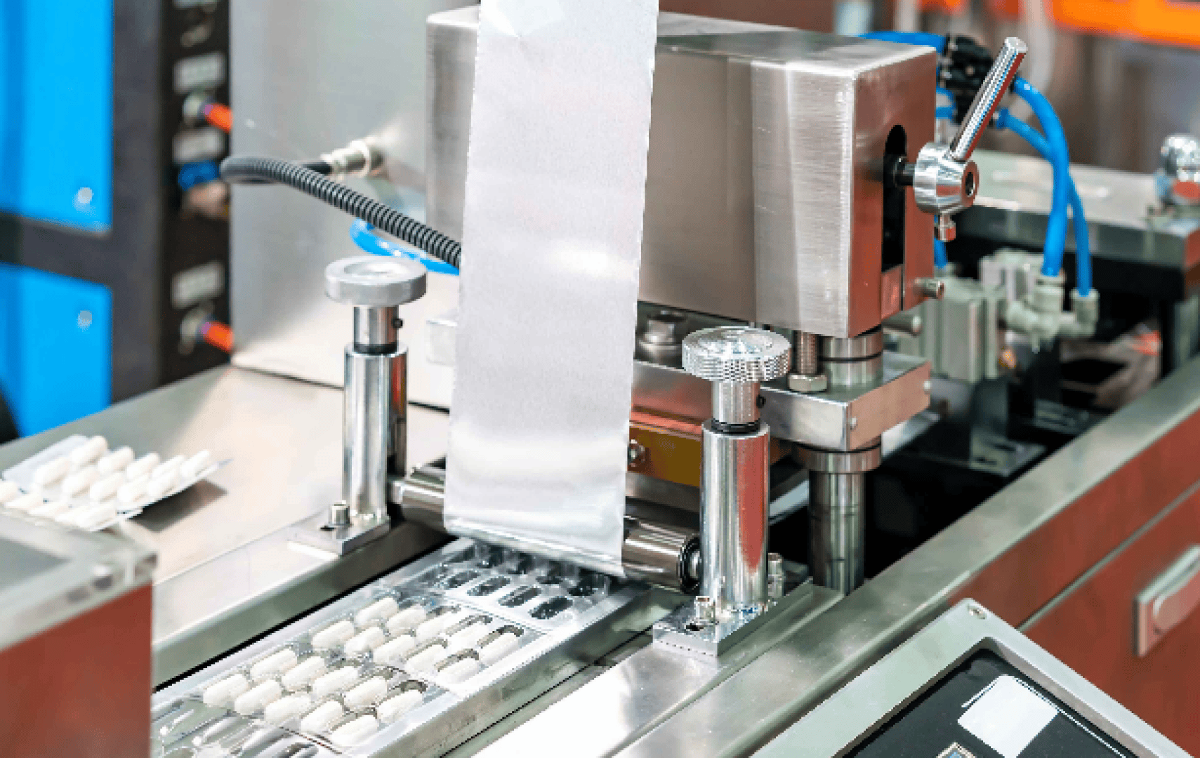 Close-up of a pharmaceutical packaging machine sealing tablets in blister packs, with metallic components, control knobs, and a strip of packaging material running through the equipment.