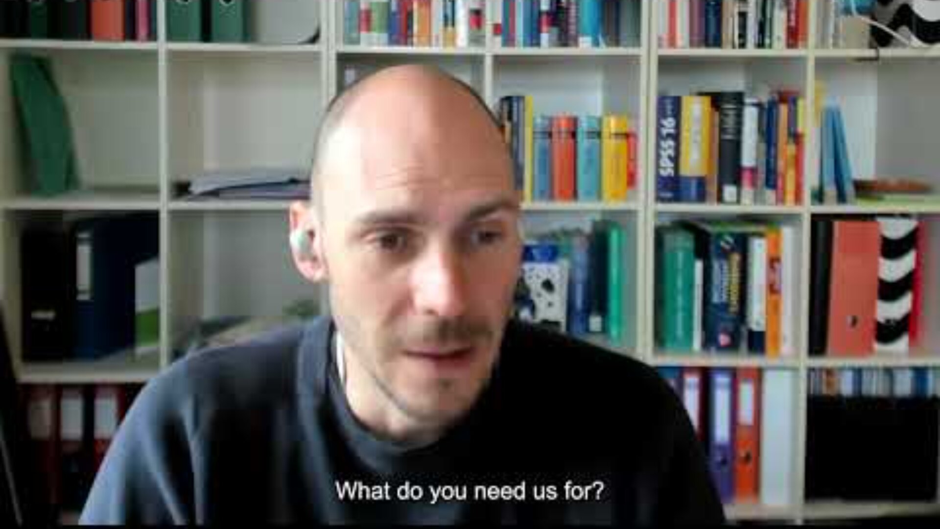 A man with earbuds sits in front of a bookshelf filled with books and binders, speaking during a video call. Subtitles at the bottom read, What do you need us for?.