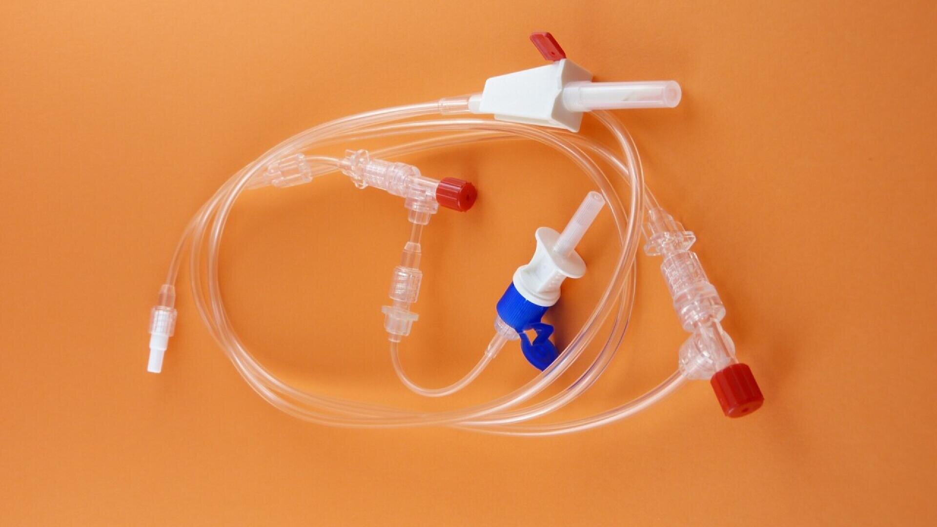 Clear medical tubing with connectors and caps in red, white, and blue, coiled on an orange background. The tubing has various ports and valves for medical use.