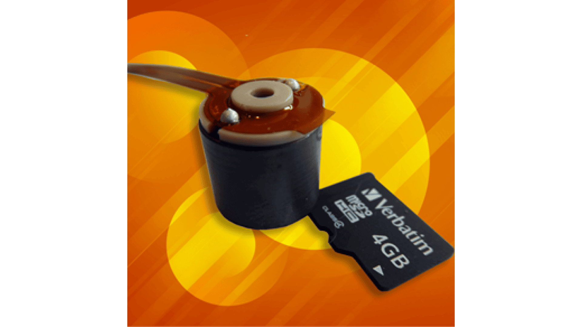 A close-up of a small black electronic component with wires next to a 4GB Verbatim microSD card on an orange and yellow abstract background.