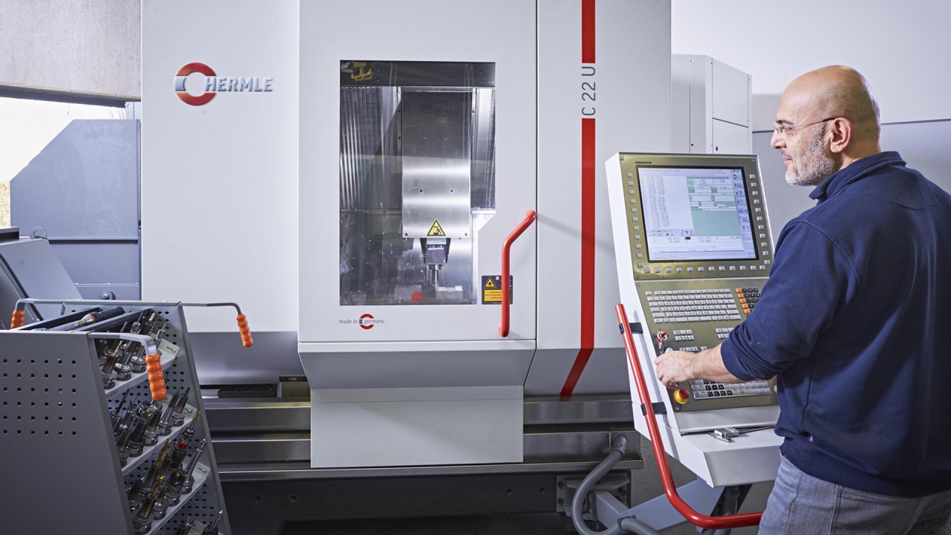 Ein Mann bedient eine CNC-Maschine über ein Bedienfeld in einer Industrieumgebung, in deren Nähe verschiedene Werkzeuge und Geräte zu sehen sind. Die Maschine ist von der Marke Hermle und mit C 22 U beschriftet.