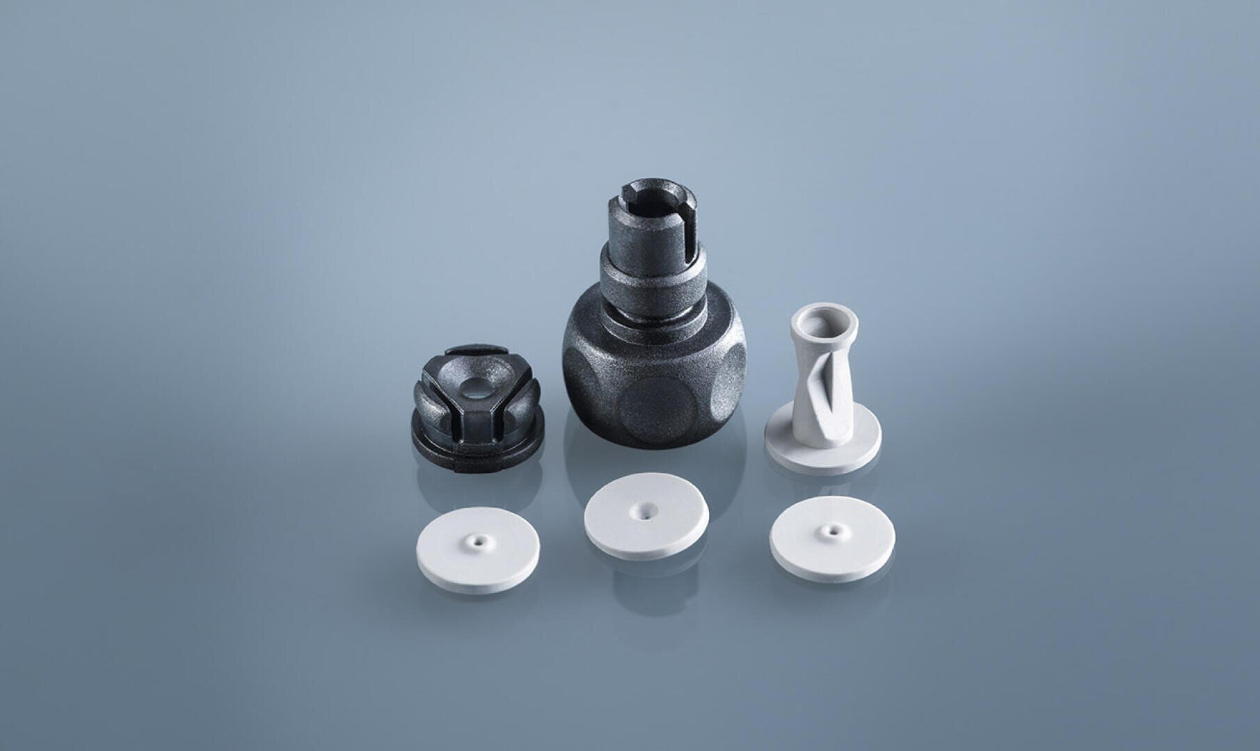 Five small, precision-engineered mechanical components are arranged on a light blue surface; three are flat, circular white pieces, one is a white cylindrical part, and one is a larger, metallic black piece with a complex shape.