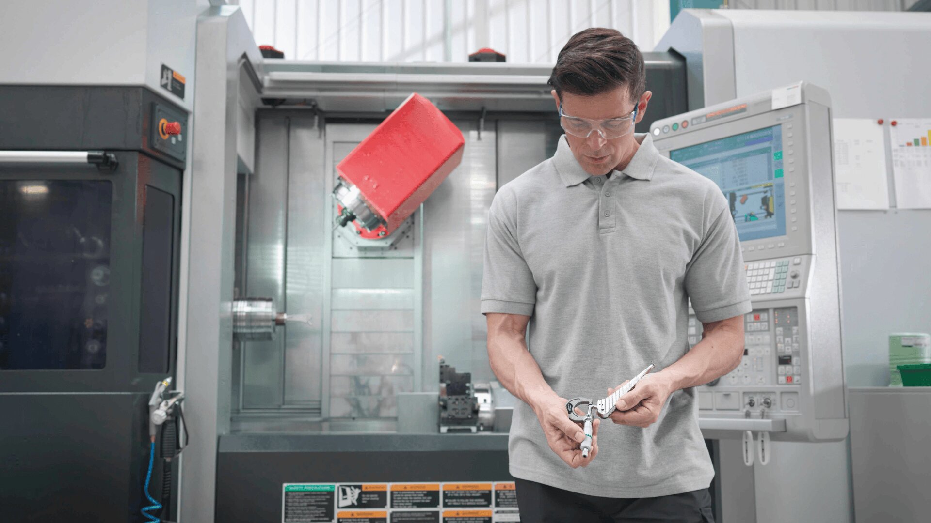 Ein Mann mit Schutzbrille prüft ein Metallteil vor einer großen CNC-Maschine in einer Industrieanlage. Hinter ihm ist im Inneren der Anlage ein rotes Maschinenteil zu sehen.